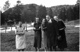 Photo of a group of people at McGraw Ranch, circa the 1930s. On the right, there is a group of 4 guests--one young man and 3 young women, dressed in coats and laughing and smiling with each other. To the left, there is a middle-aged woman who appears to be a ranch staff member, as she is dressed in a plain dress covered by an apron. She has her arms crossed, with a reserved smile. In the background, there are fences and an outhouse, and mountains in the background.