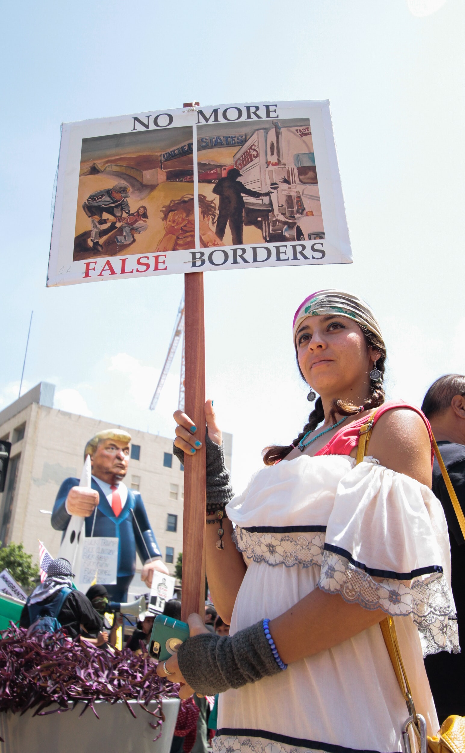 Photo of a young woman taking part in a peaceful protest, demonstrating the Trump Administration's border and Immigration policies. She is carrying a protest sign that says "No more False Borders." The sign shows an immigration officer throwing a person to the ground on one side, and on the other side the image shows semi truck after semi truck carrying guns going right past border officials.