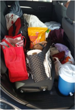 Photo of grocery bags on the back seat of a car. The bags are full of snacks and supplies for a road trip, and a bag of popcorn as well as a roll of paper towels are visible in the tops of two of the reusable canvas grocery bags. A blue insulated cooler, possibly of water, sits on the floor in front of the seat.