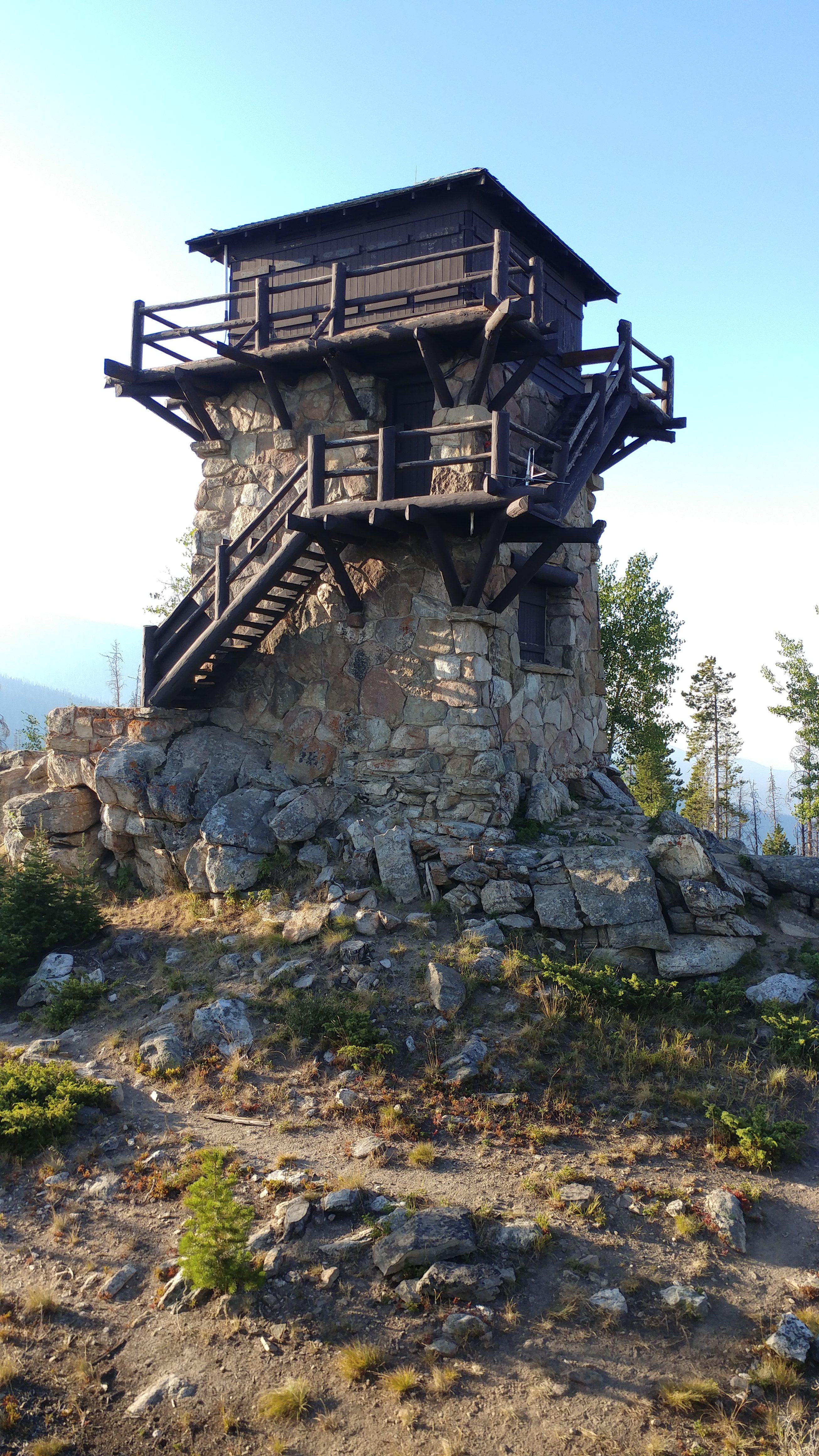 Photo of the stone and wood structure that is the Shadow Mountain Fire Lookout tower. It is a tall structure, square in shape, with a wooden deck and room that sits atop a stone base.