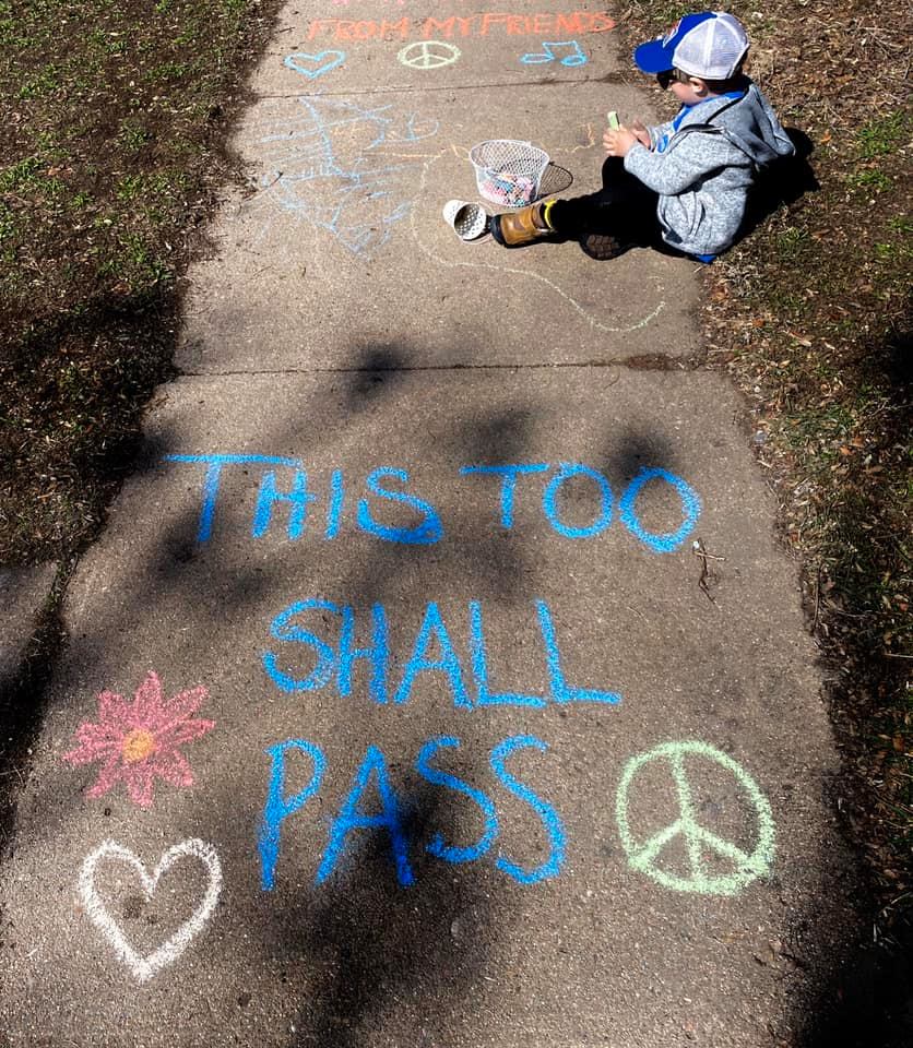 Photo of a city sidewalk, flanked by grass on each side. A child sits on the edge of the grass toward the top of the photo, and they are holding sidewalk chalks. A bucket of chalks is on the ground next to their foot, and they are wearing jeans, a sweatshirt, and a hat. The sidewalk is decorated with hearts and flowers in yellows, blues, and pinks, along with yellow peace symbols. In large blue letters are the words "This too shall pass."