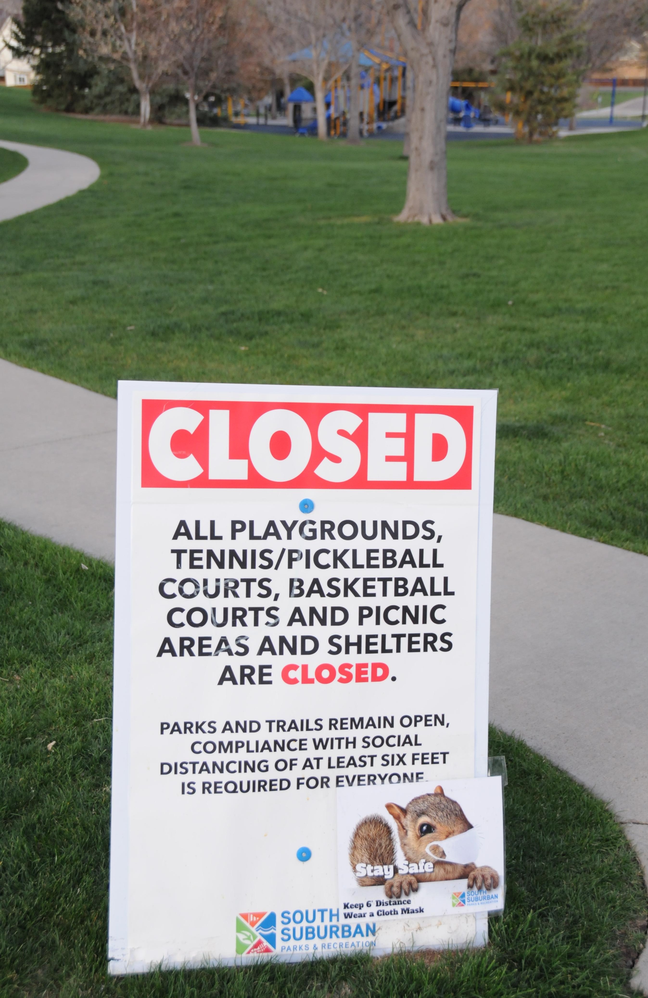 Photo of a sign posted in a park, indicating that the park has been closed due to pandemic protocols. It says," Closed: All playgrounds, tennis/pickleball courts, basketball courts, and picnic areas and shelters are closed. Parks and trails remain open, compliance with social distancing of at least 6 feet is required for everyone." There is a photo of a squirrel wearing a face mask at the bottom of the sign.
