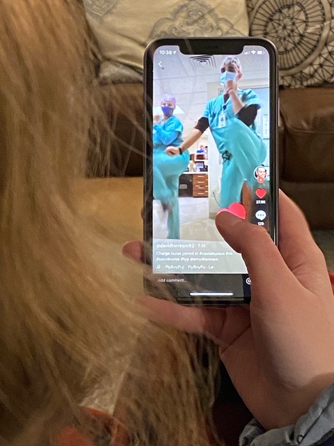 Photo of a person holding their cell phone and watching medical personnel dance. The personnel are dressed in scrubs and facial masks. The video is being played on the TikTok app.