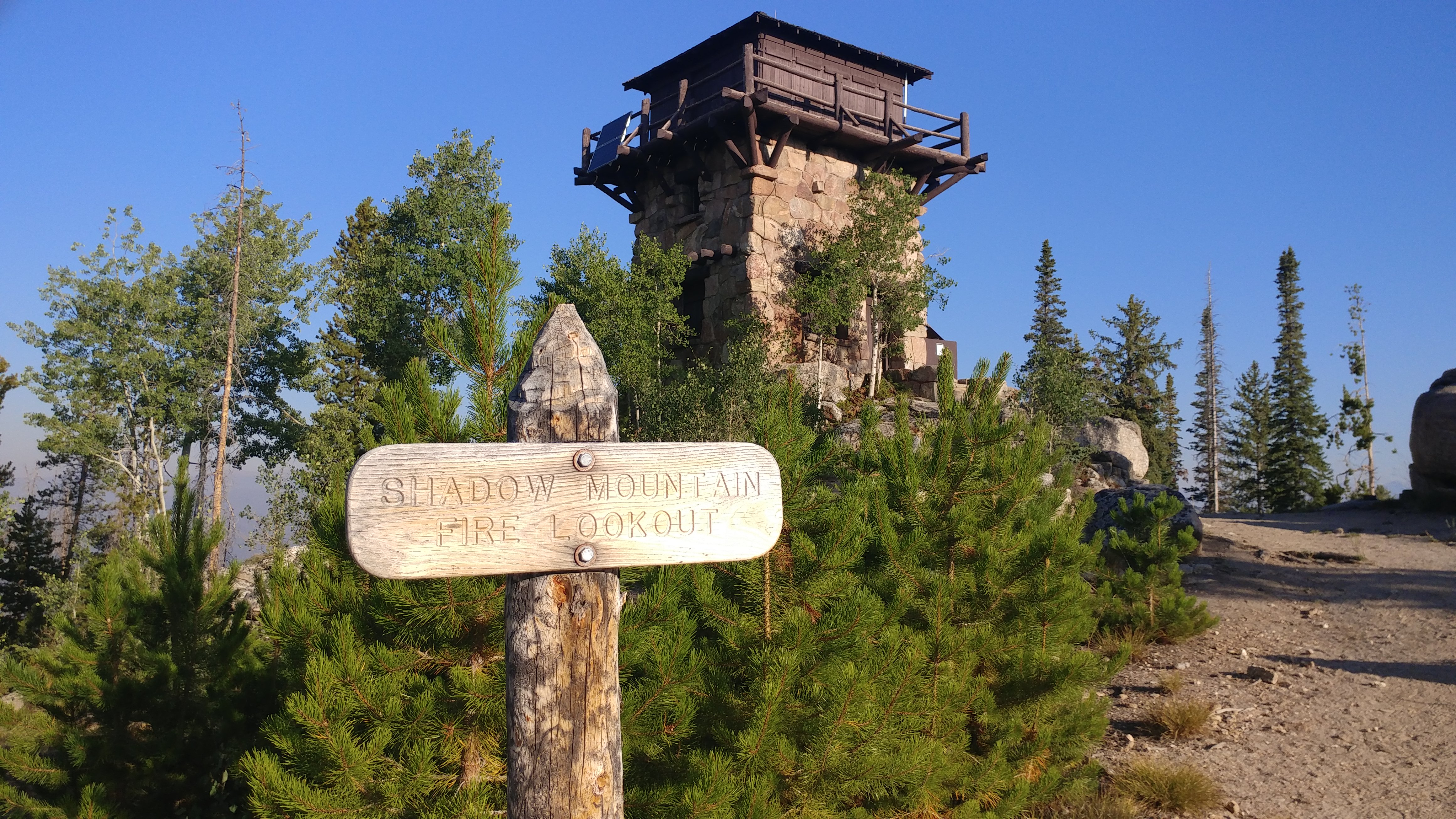 Photo of the sign posted for the Shadow Mountain Fire Lookout. The sign itself is handmade made of a log about five inches in diameter, which serves as the post for the sign and has been whittled into a point at the top. Bolted to the post is a flat wooden plaque in which the name "Shadow Mountain Fire Lookout" has been carved by machine in block letter.  Along the trail in the background stands the fire tower.