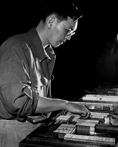 Photo of Japanese American male using small metal letters to lay out type in order to print something on paper, such as a newspaper