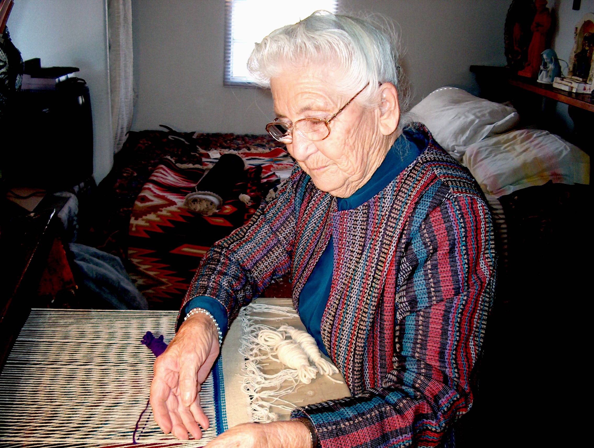 Photo of an older woman with white hair and eyeglasses, looking down at her weaving loom as she works. She is carefully lifting some of the threads as she weaves a piece of fabric that is white with a bright turquoise horizontal stripe.