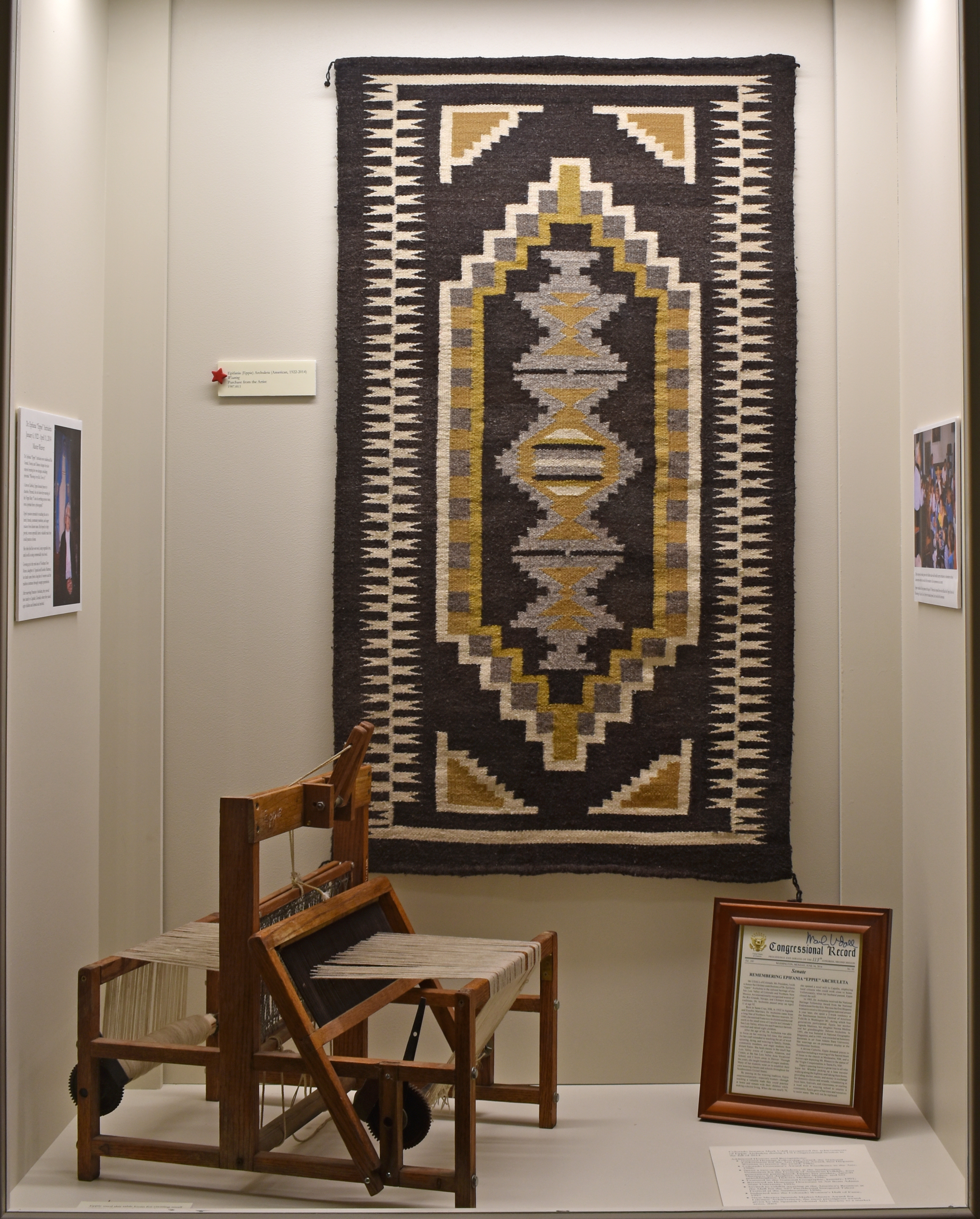 Photo of Eppie Archuleta’s weaving on display in the Adams State University Luther Bean Museum. The display includes a copy of the Congressional Record honoring Eppie for her lifetime contributions. It also displays a table loom made by Levi Medina and donated by Levi and Norma Medina. 