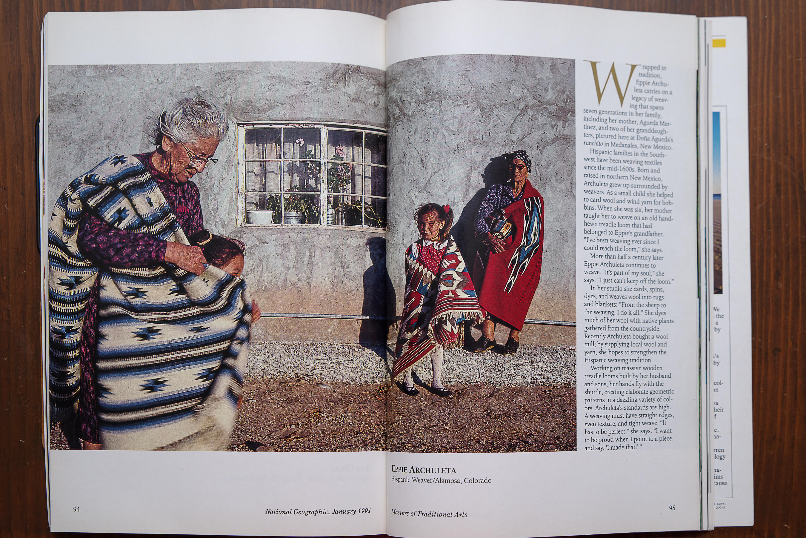 Photo of a magazine, opened to show a large photo across two pages. The photo is of Eppie Archuleta standing with her granddaughter, a woven blanket over Eppie's right shoulder and wrapping around the young girl in front of her. They are standing outside of a white-walled building with a paned window, near where another older woman and young girl both stand, with blankets of red and turquoise wrapped around them.
