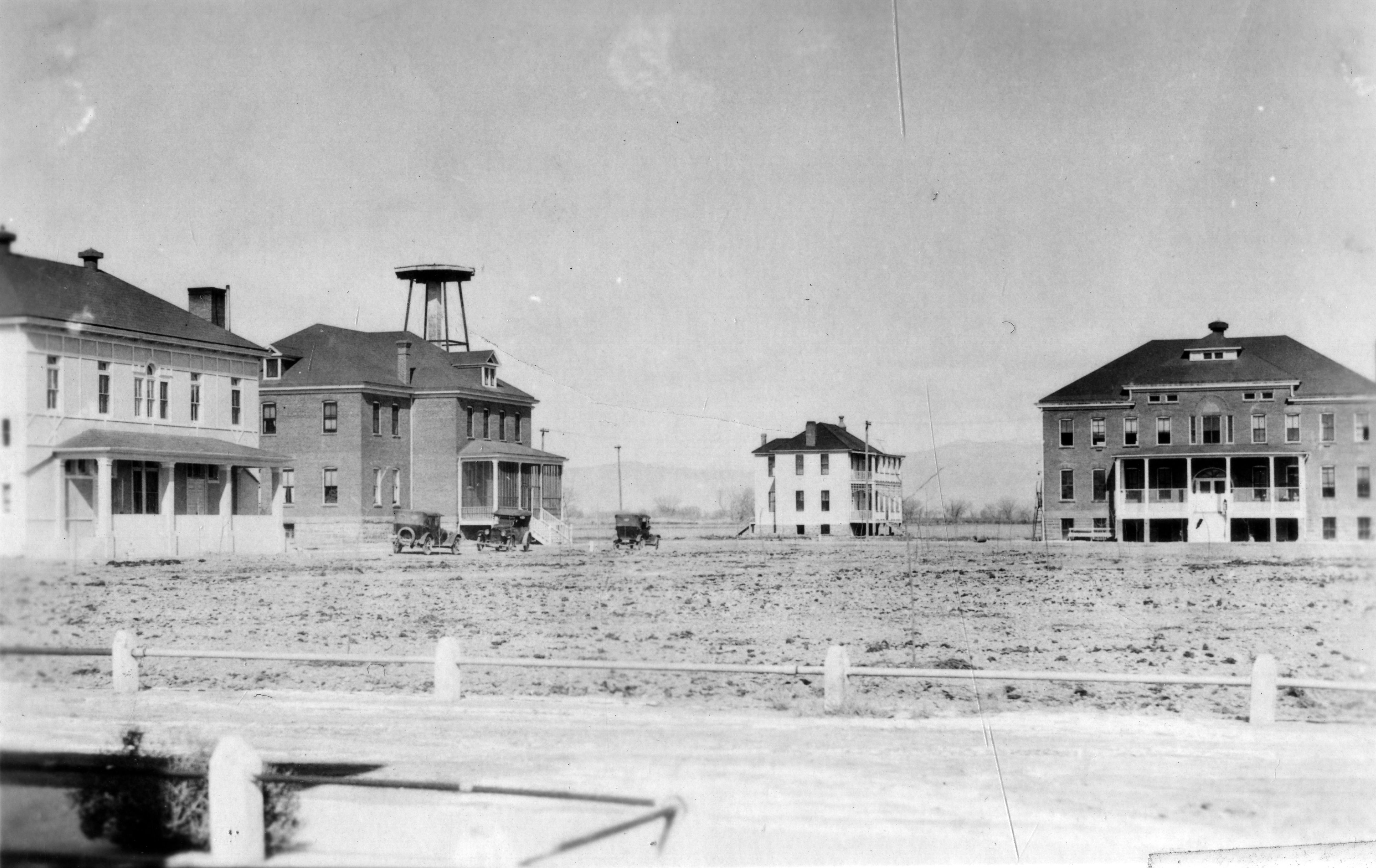 Photo of four buildings sitting on a barren piece of land. in the foreground, part of a dirt road can be seen. There are 3 vehicles parked in front of two of the buildings on the left side of the image. In the far distance, the hazy silouette of mountains can be seen. There are no people immediately visible in this historic photograph.