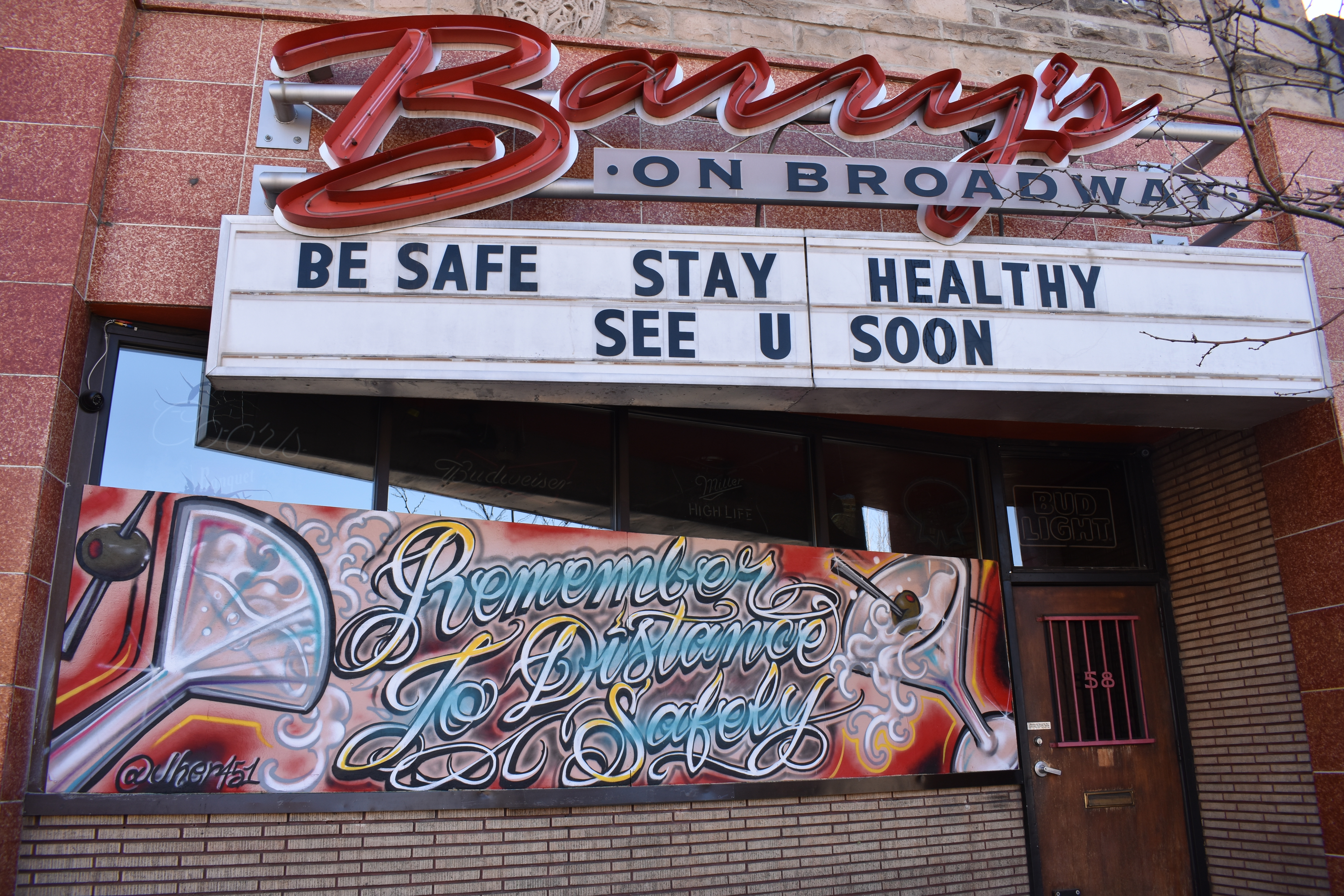 A photo of a closed bar along Broadway in Denver, taken in April of 2020. The marquee sign above the door at Barry's on Broadway says "Be Safe - Stay Healthy - See U Soon." The owners have boarded up their windows during the stay at home order, which artists have decorated with images of martinis and the words, "Remember to Distance Safely."