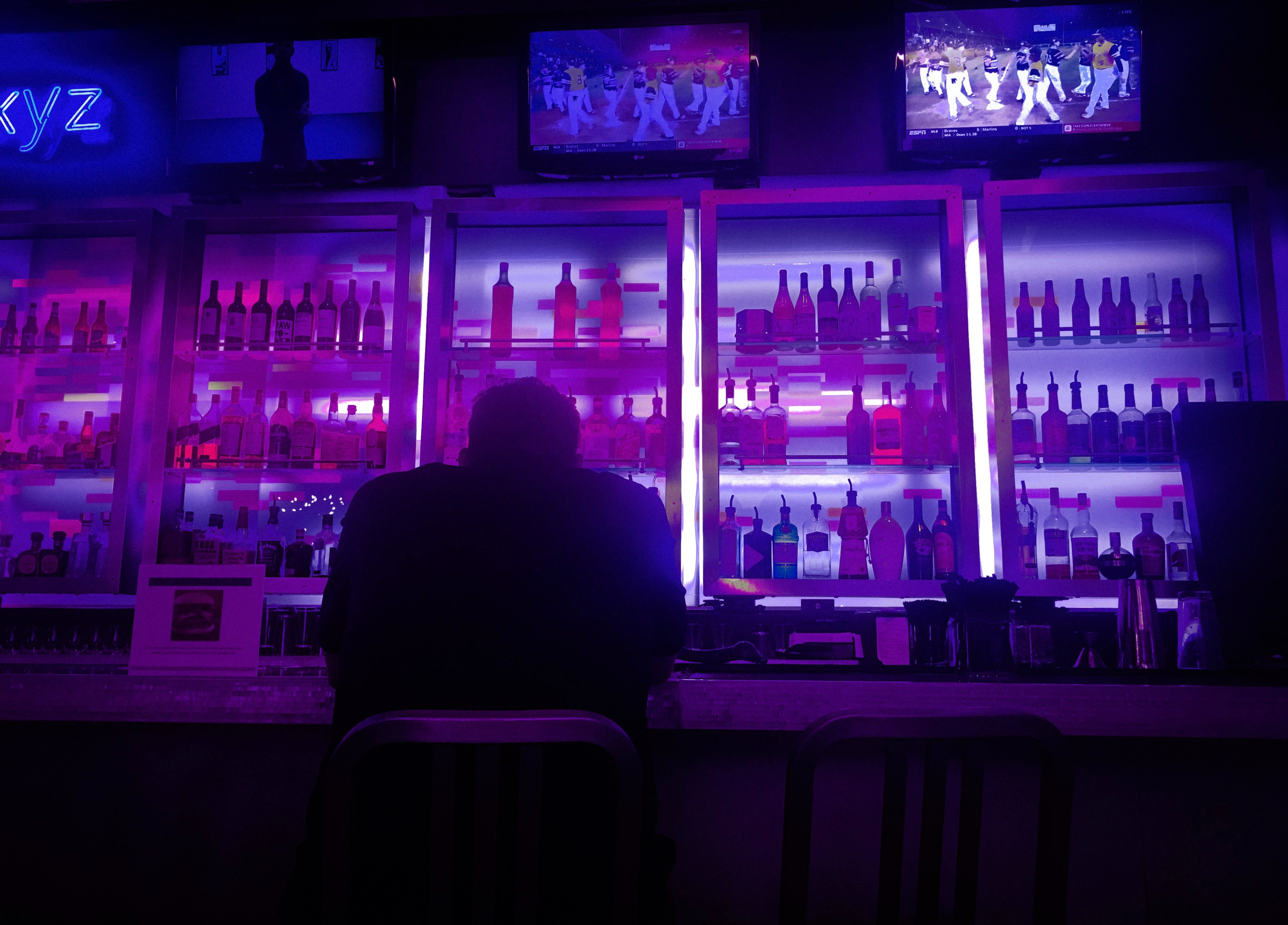 Photo of a person sitting alone at a bar. Their back is toward the camera, and they are hunched over. The bar in front of them is backlit with purple lighting, giving off an interesting glow and showcasing the glass shelves full of alcohol bottles behind the bar.