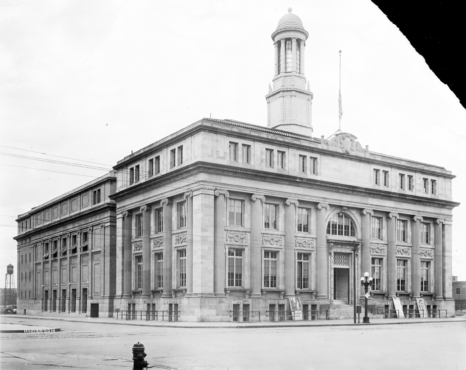 Photo of a grand building constructed of a light colored stone brick. Tall columns line the front and side of the building, between which are paned windows from the ground and first floors. A large paned semi-circular window sits above the front door in the center of the building, and a tall tower emerges directly above that from the roof. An American flag flies at half-staff on the flagpole affixed to the roof above the front door in this historic black and white photo.