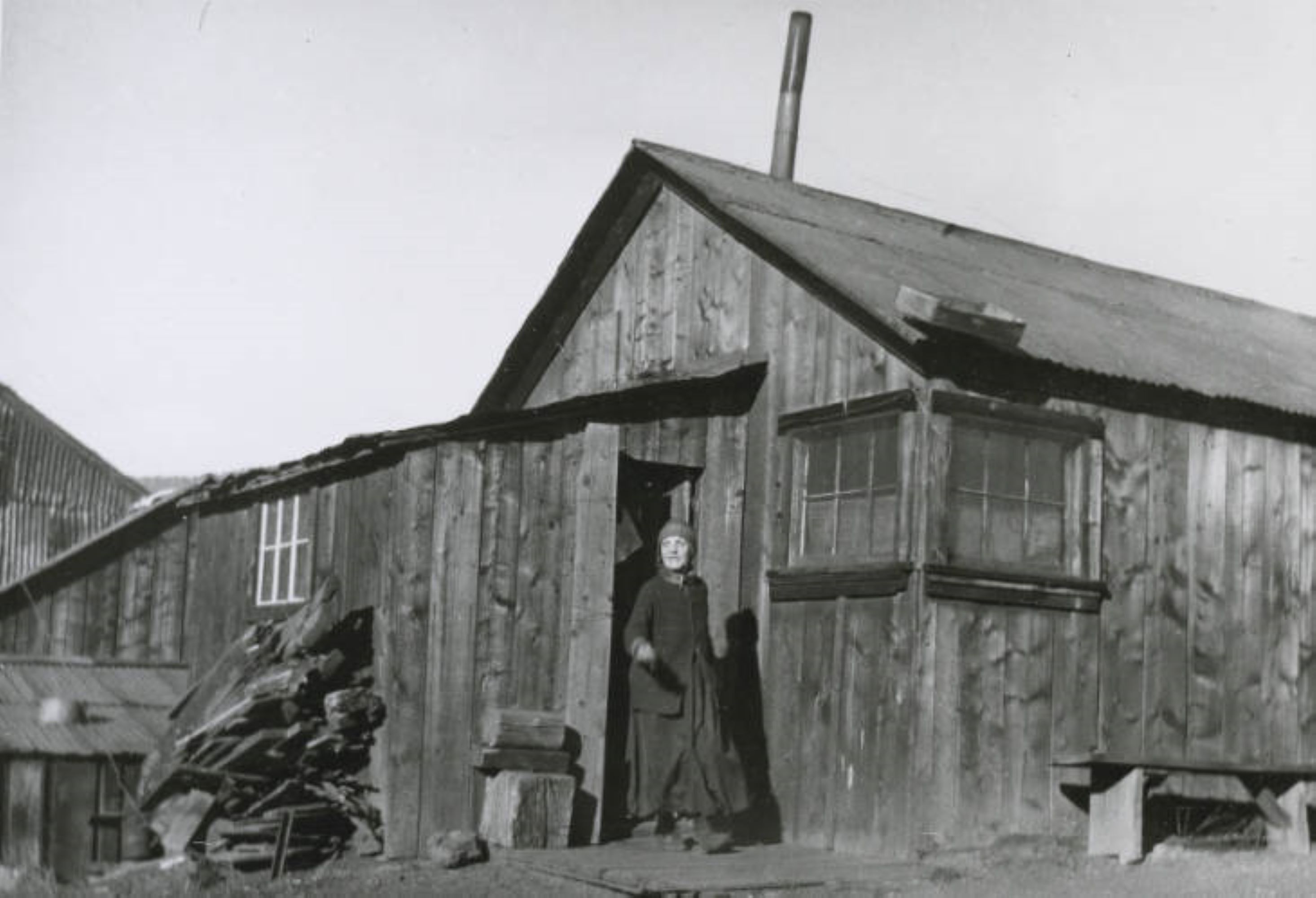 Photo of a woman in layers of dark clothing, boots, and a cold weather stocking cap, walking out of the door of a wooden building. The building has just a few small windows and a metal roof with a metal chimney pipe coming from the top of the roof. There is a stack of firewood around the corner from the door, and a bench around the corner to the left of the door. 