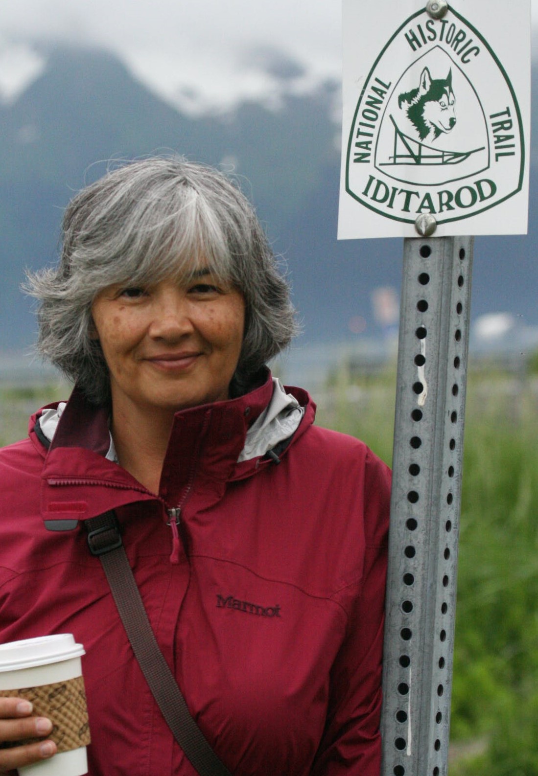 Photo of author Susan Harness. She is standing next to a road sign that reads "National Historic Trail" IDITAROD." She is smiling, and holding a beverage in a coffee takeout cup. She is wearing a zipped bright red outdoor jacket that has a hood attached, and a cross-body bag of some kind. She has short, salt-and-pepper colored hair with long bangs that hang to her eyebrows. In the distance behind her are tall, snow-capped mountains.