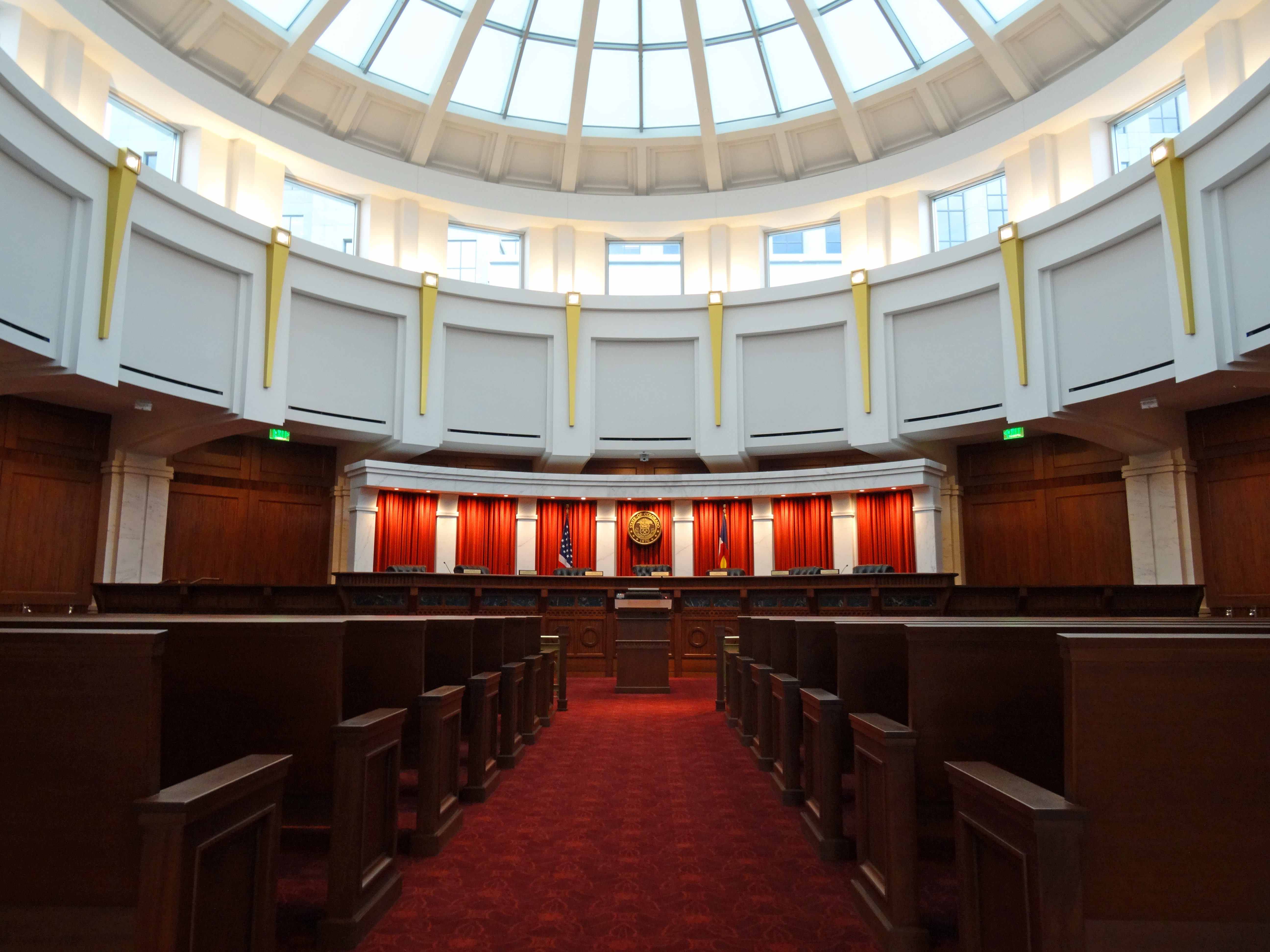 Photo of the inside of the Colorado Supreme Courtroom. The red-carpeted aisle passes by about 7 rows of dark wooden benches, as the Bench looms at the back of the room. There are 7 sets of red curtains against a white cabinet, and in the center set of red curtains hangs the seal of the State of Colorado. The round room is circled by a white balcony with gold trim, and the ceiling is a set of windows creating a large circular ceiling of glass.