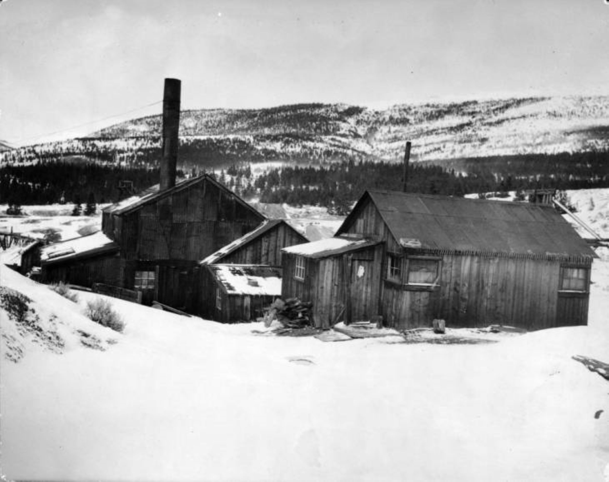 Photo of the historic Matchless Mine. Wooden building stand in the center of the black and white image, in which snow has fallen and is covering the ground around the buildings as well as dusting the mountains visible in the background. The buildings have few windows, and two of them have metal chimney pipes standing up in the air. Icicles hang from the metal roofs. There are no people visible.