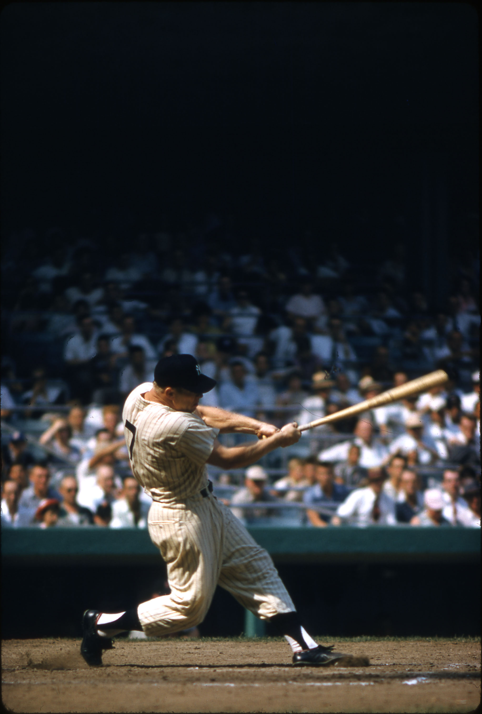 Photo of baseball great Mickey Mantle, swinging the bat at an oncoming pitch during a baseball game. He is leaning into the swing and swinging the bat to his left in this photo. He is wearing a dark baseball cap, and a white pinstriped uniform with black shoes. On his back is the number 7. In the background, crowds sit in the stadium stands watching the game.
