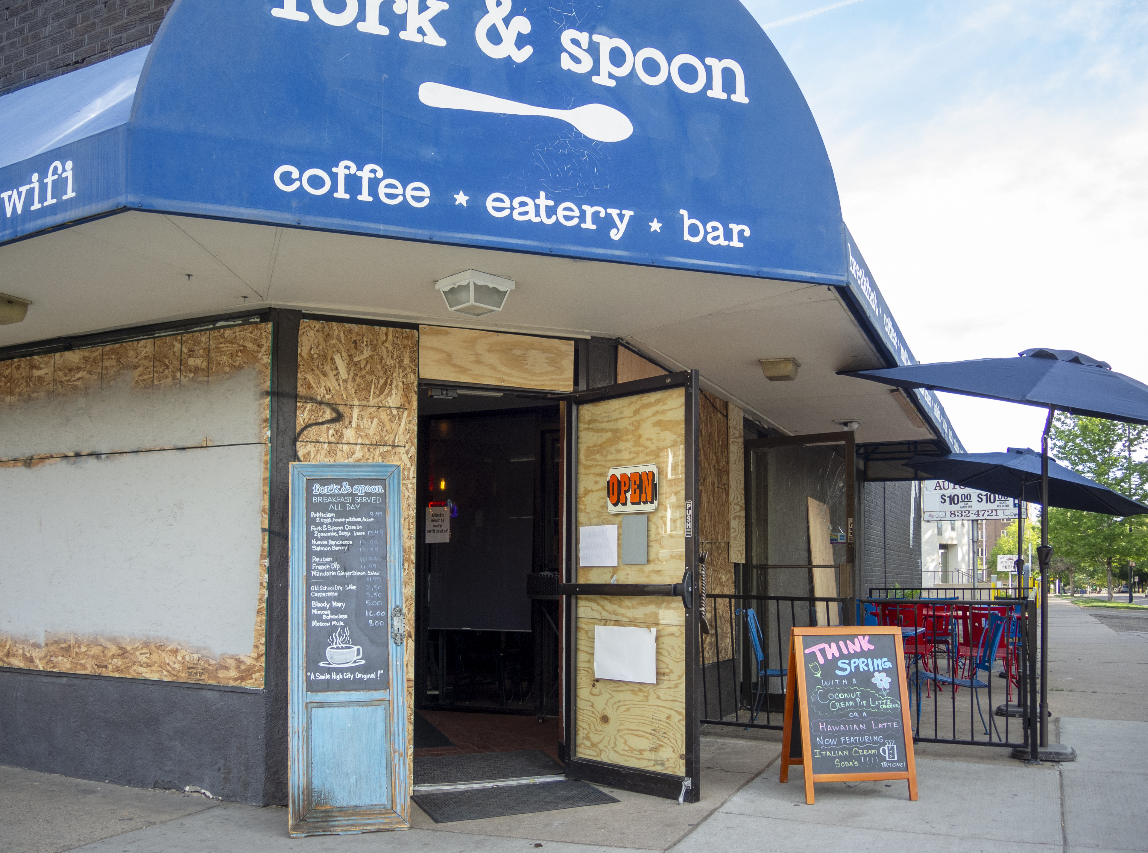 Photo of an eatery in Denver called fork & spoon. Their front door is open, but their windows are boarded up as they had been closed due to the pandemic lockdown. There are a few tables set up on the sidewalk for outdoor dining, and a sign posted inside the front door says "Masks must be worn until seated!"