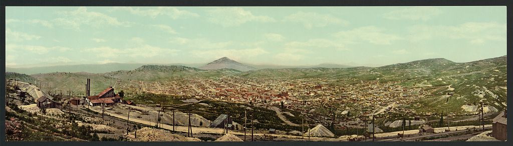 Photo in panoramic view of the town of Cripple Creek, Colorado in the year 1900. Mountains are seen in the far distance, and in the foreground mining operations are visible. The town stretches out in the valley.