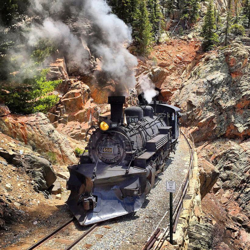 Photo of the 489 steam locomotive of the Cumbres and Toltec Scenic Railroad, having emerged from a tunnel and traveling around a slight curve, toward the viewer. It is a sunny day, and green trees dot the red and grey rocks of the mountains on either side of the black engine.