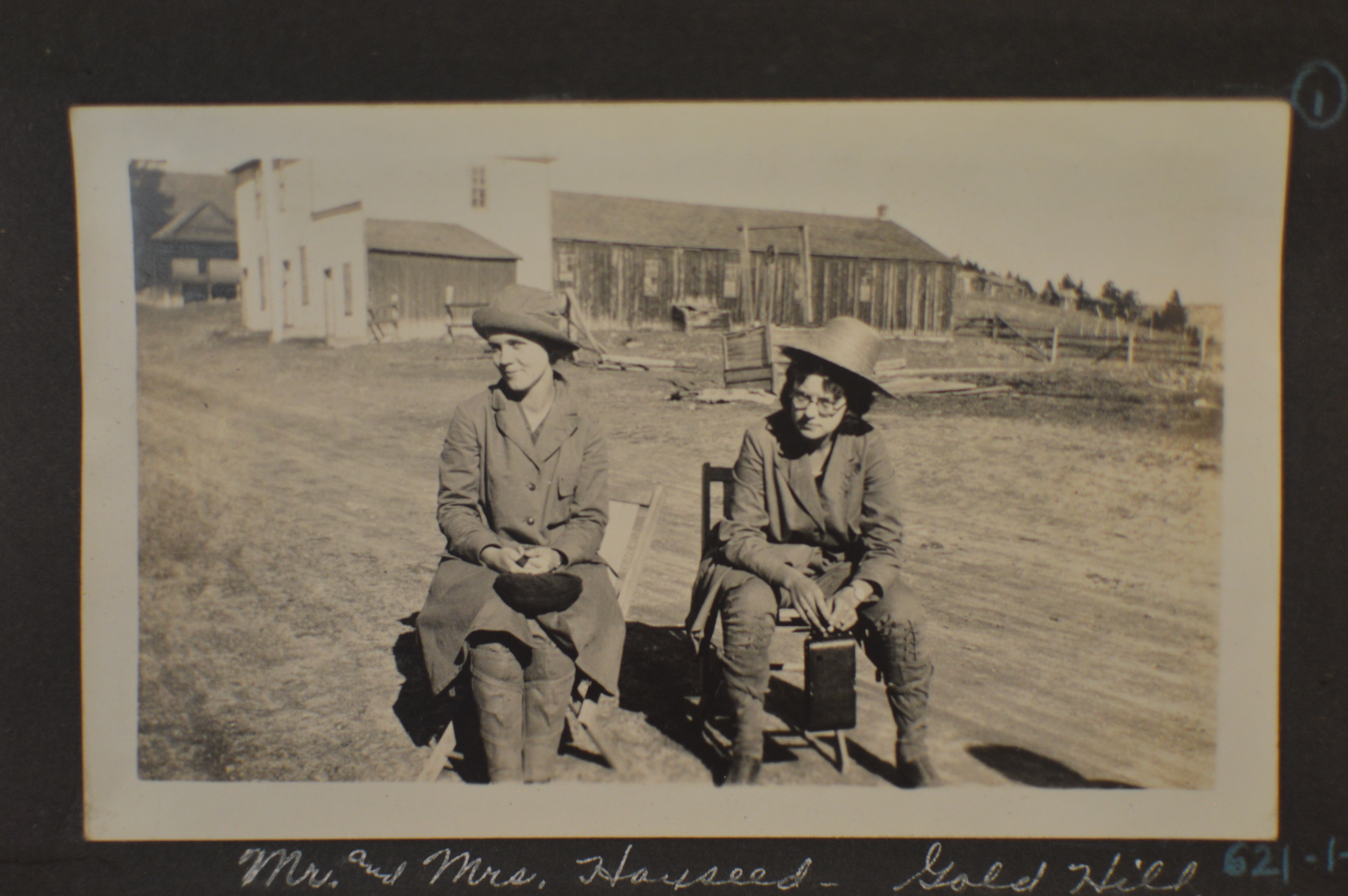 Photo of two women outdoors, dressed in tall boots, long coats, hats. The caption, handwritten at the bottom of the old photograph, says, "Mr. and Mrs. Hayseed--Gold Hill." The young women are sitting on folding chairs; the woman on the left holds a handbag on her lap and smiles at the photographer. The woman on the right is dressed in a more masculine style of clothes for the day, including trousers and a unisex-style hat. She is looking directly at the camera, with a slightly more serious expression.