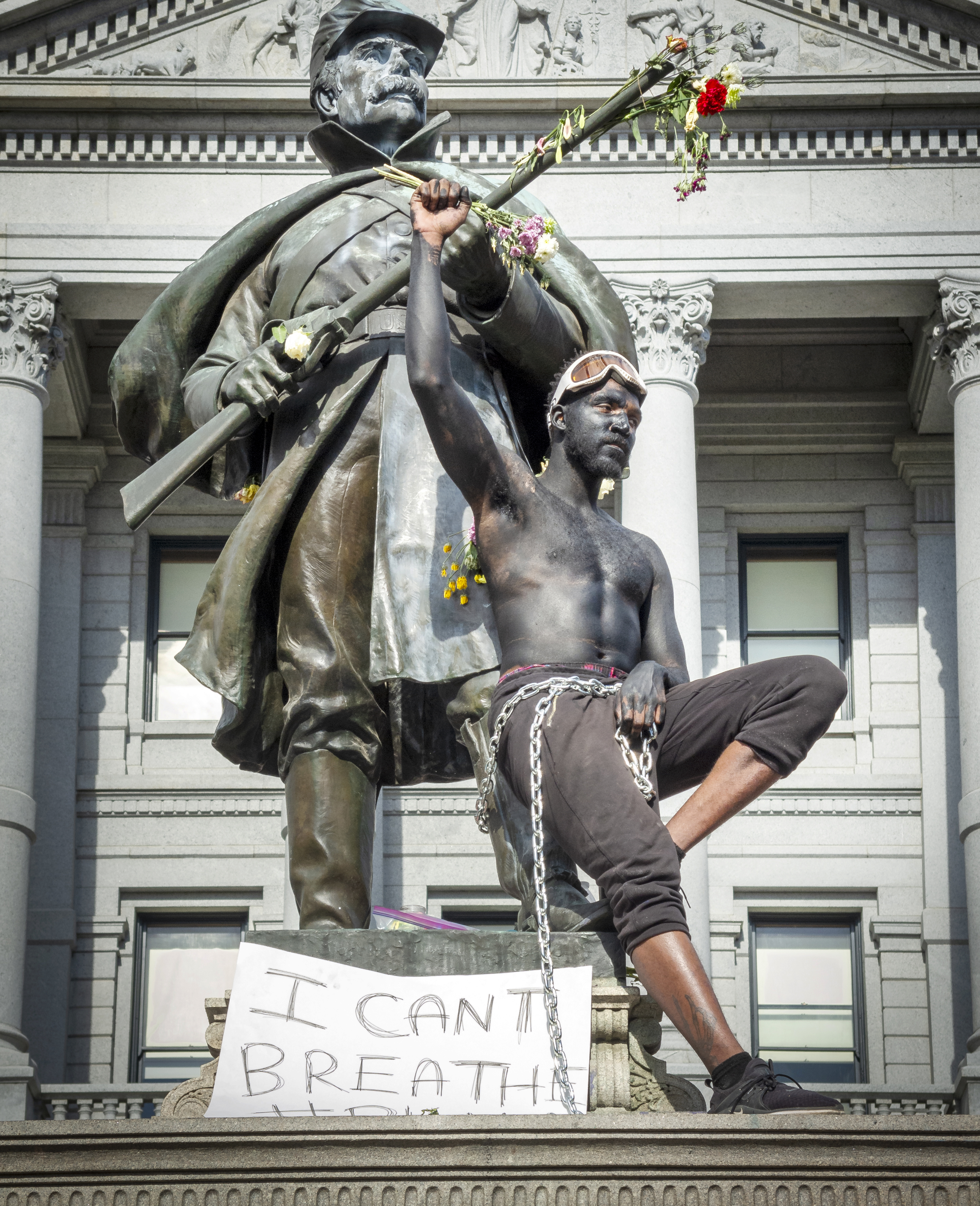 Photo of a Black Lives Matter protestor with a sign that says, "I can't breathe." He is seated at the base of a statue of a Civil War soldier. the protestor has painted himself to look bronzed like the statue, and he wears pants, chains around his waist, shoes, and white ski goggles that sit atop the protestor's head. His right fist is held high in the air. The statue of the soldier is holding a rifle, and flowers have been stuck into the end of the firearm.