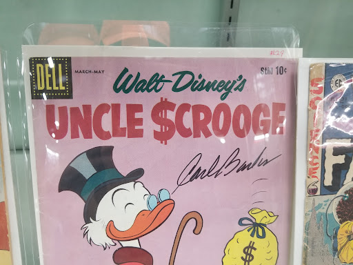 Photo of a comic book in a rack with other books. The book is in a clear plastic sleeve. The cover is a pink background, with a title across the top of the book that says "Walt Disney's Uncle Scrooge." The top part of an image of the cartoon character Uncle Scrooge is seen, wearing a top hat, spectacles on his beak,  and smiling. A yellow bag with a dollar sign on it is next to him.