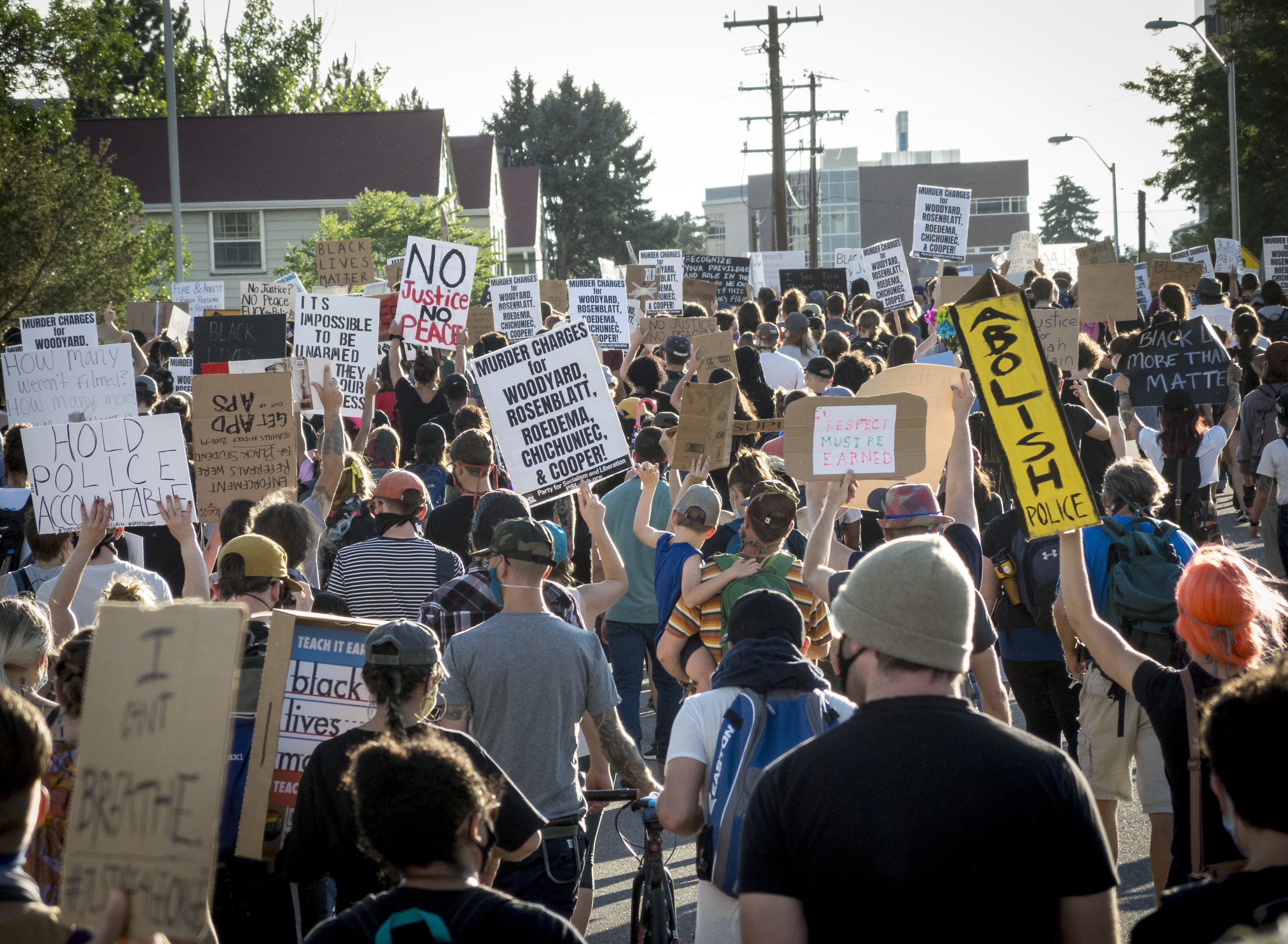 Photo of protestors attending a march and protest, walking down the street and holding their empty hands into the air, while some people are holding up signs. One sign reads, "Hold Police Accountable," while another reads, "I Can't Breathe." It is a warm sunny day, as the crowd wears warm weather clothing, any many among the crowd wear sunglasses, hats, and face masks.