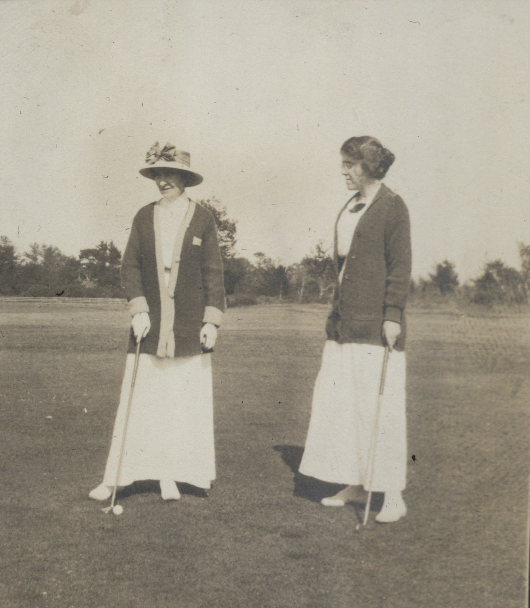Photo of two women in the early 1900s, playing golf. They are both wearing long white skirts, with white shoes, white blouses, and cardigans that are hip-length and are buttoned in front near the waist. The woman on the left is wearing a sun hat with a wide brim and big bow in front, while the woman on the right wears no hat, her hair pulled back into a bun. The are each leaning slightly on their golf clubs which are perpendicular to the ground. The woman on the right appears to be lining up her ball.