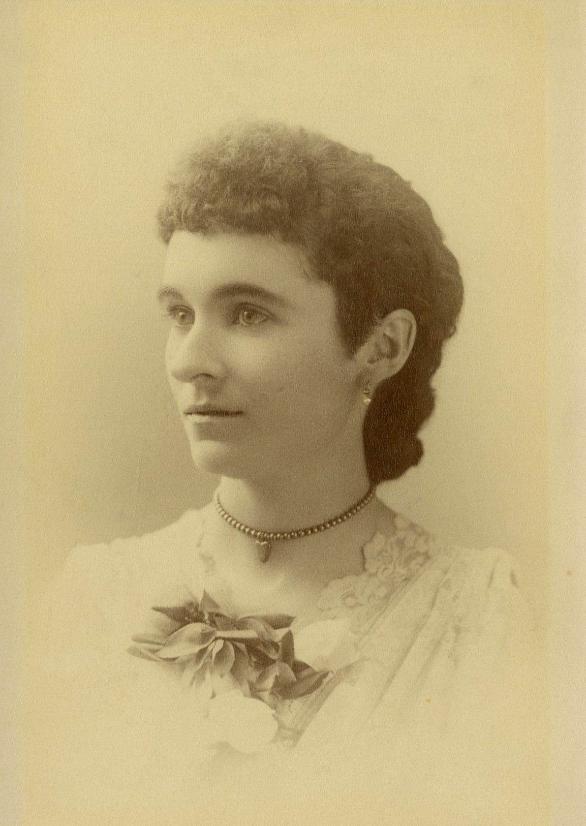 Photo portrait of a young woman, a three-quarters view of her face and shoulder, she is looking off to the left. She is wearing a white lace V-neck blouse with a flower corsage pinned in front. She has short dark hair that looks thick and wavy, with very short bangs across her forehead. She wears small earrings of a dangling pearl, and a beaded choker necklace that has a heart-shaped pendant.