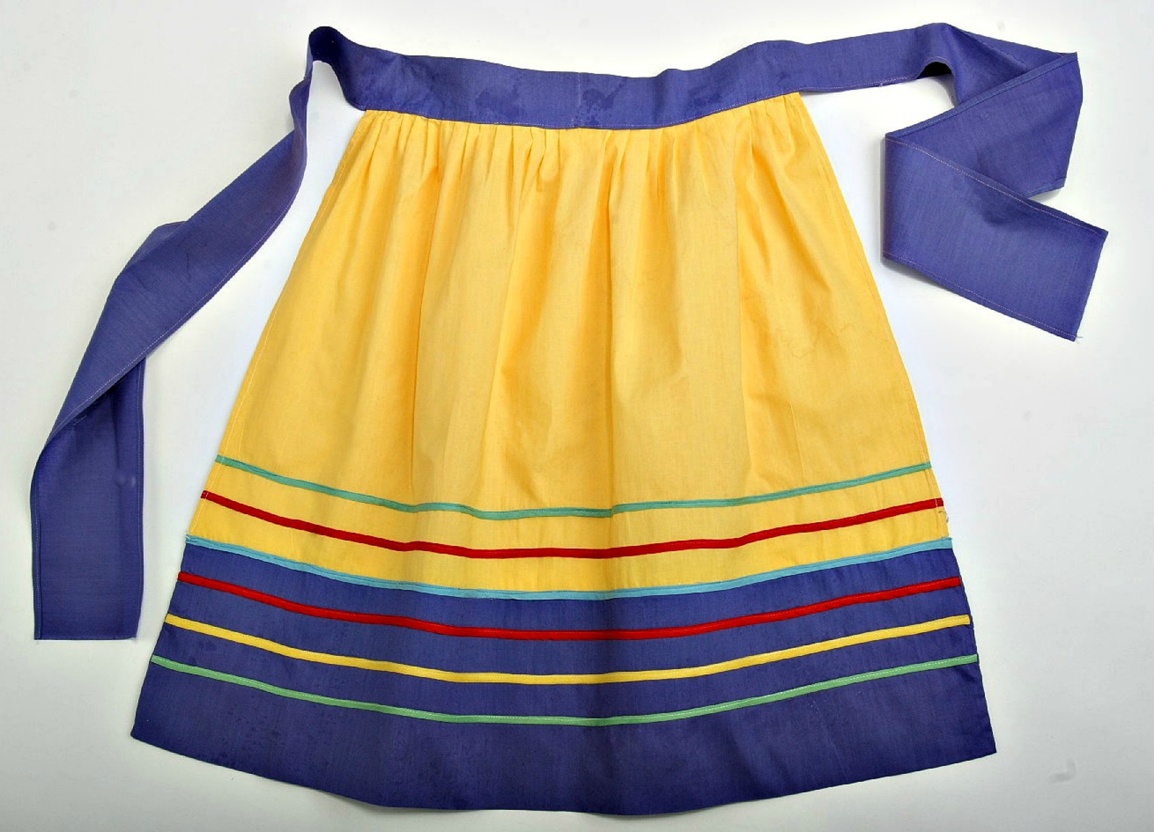 Photo of an apron. It is bright yellow, with a wide blue waistband and bottom border. Along the bottom of the apron are thin stripes of green, yellow, and red, created by bias tape that has been stitched to the apron.