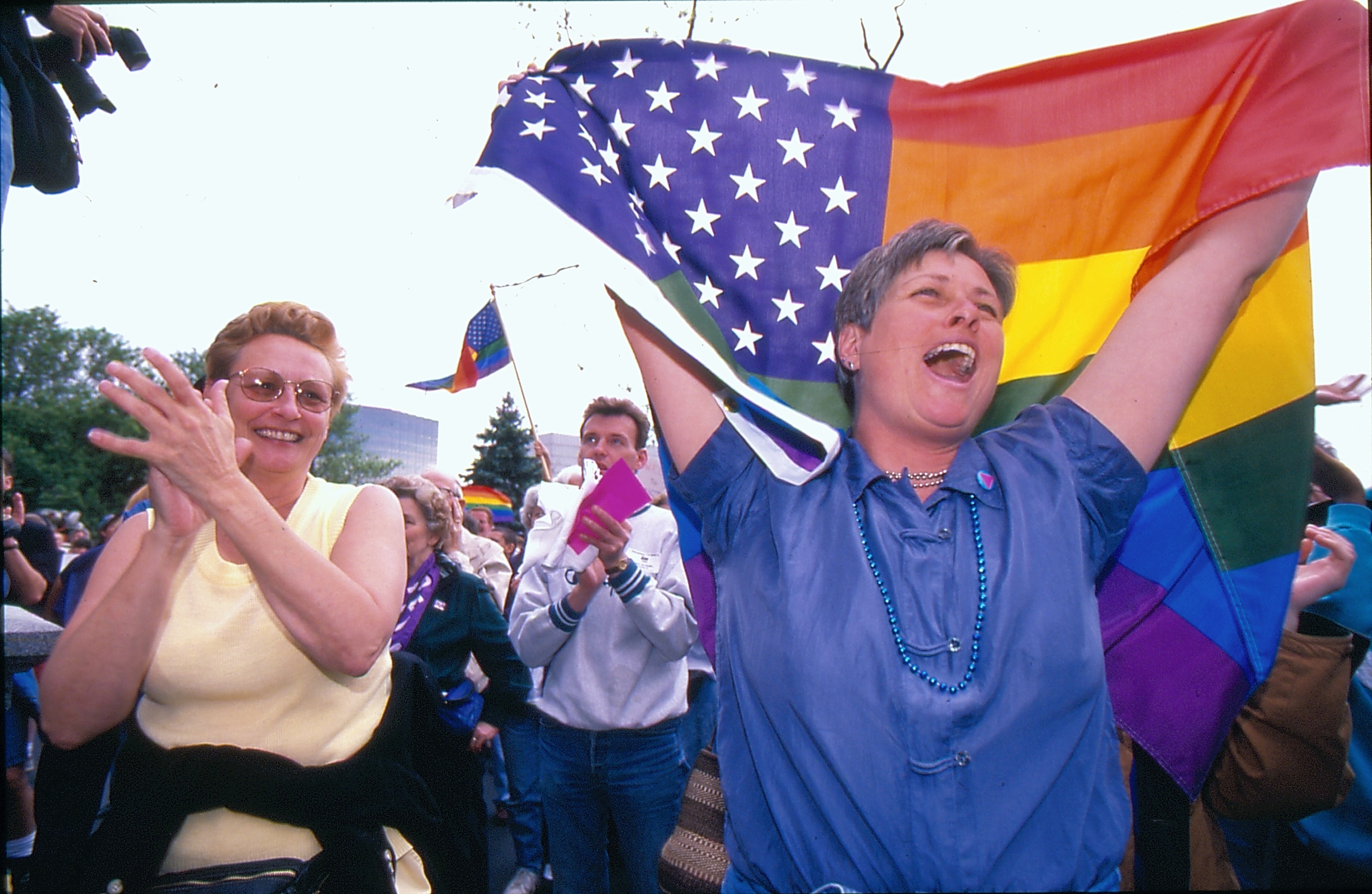 Photo of a crowd of people celebrating the US Supreme Court's decision to strike down Colorado's Amendment 2. People in the crown are smiling, clapping, shouting, and waving rainbow flags high into the air.