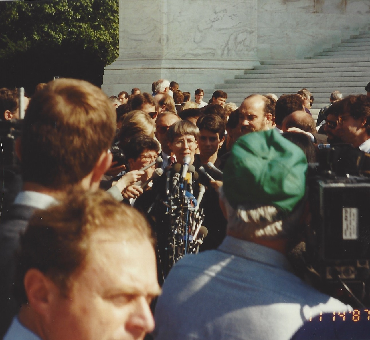 Photo of Jean Dubofsky giving a press conference. She is speaking in front of a tower of various microphones, surrounded by a large crowd of press people listening to her speak.