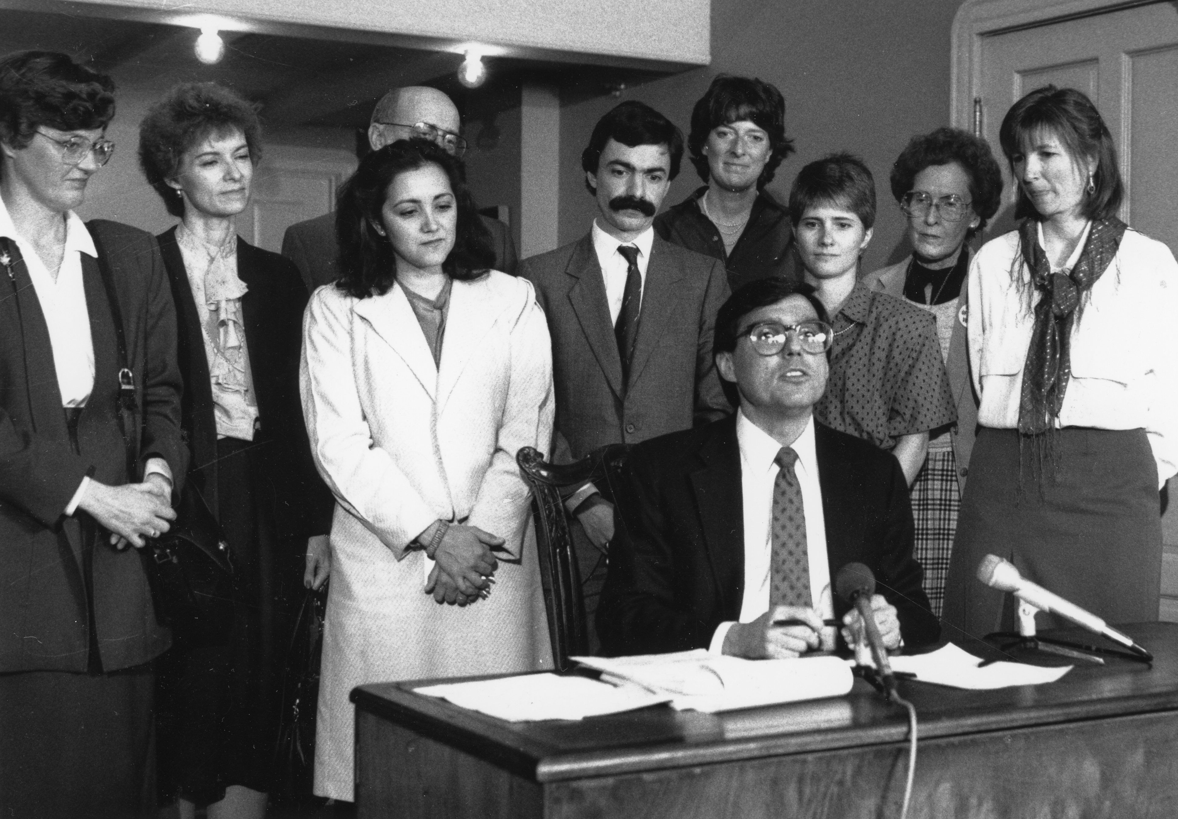 Photo of Mayor Federico Pena, sitting at a desk and talking to the media. There are papers on the desk as well as two microphones propped on the desktop and pointed toward the Mayor. He is surrounded by 9 other women and men who are there to observe the signing.