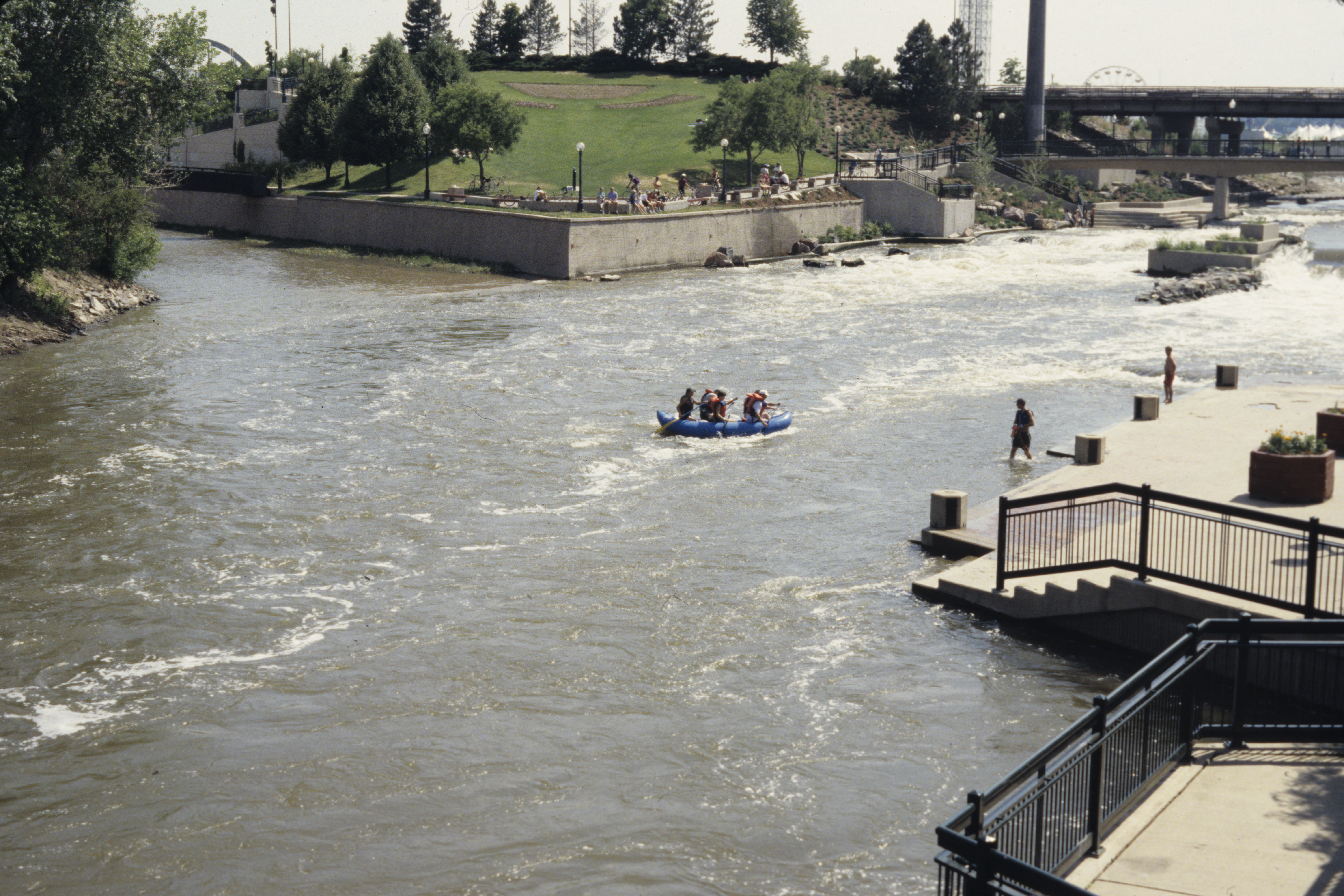 Photo of the point where two waterways meet at Confluence Park in Lower Downtown Denver. There are people floating on a blue raft in the water, while onlookers from the banks watch. 