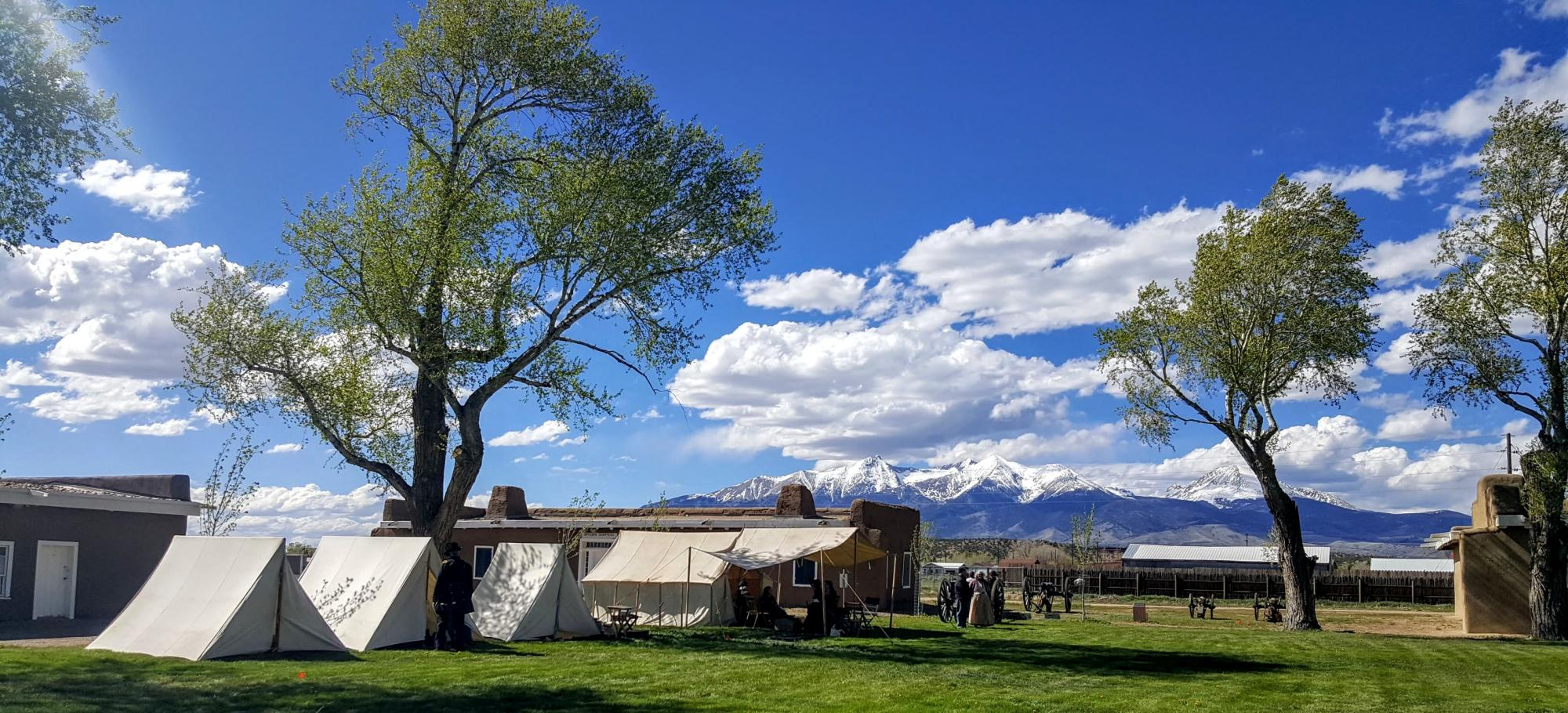 Photo of the Fort Garland museum on a sunny day, white fluffy clouds float across the brilliant blue sky. The grass is green between the low museum buildings, and a few tents have been erected on the grounds. Four cannons are toward the back of the grounds, and it appears to be a re-enactment day at the museum. Snow-capped mountains rise in the distance, touching the sky.
