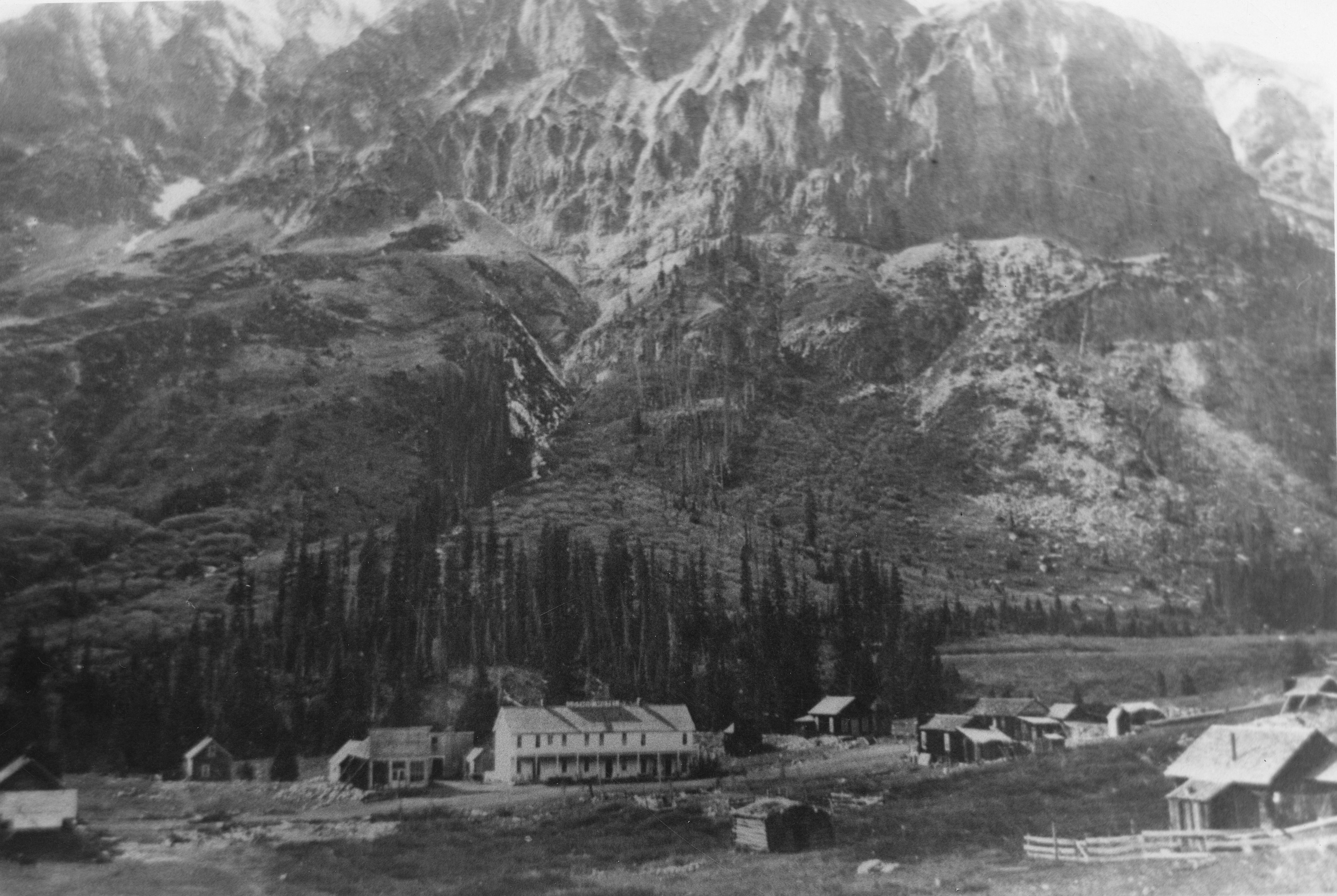 Photo of the town of Gothic, Colorado, dating from the 1880s. Buildings stand at the foot of a rocky mountain which has been dusted with snow. Most of the buildings are small one-story cabins, and one building in the center of the photo is a large white two-story building.