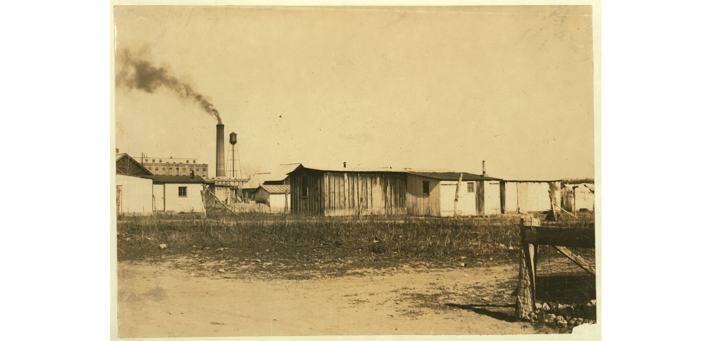 Photo of factory buildings. Many of the buildings are small one-story structures, while in the distance multi-story large buildings and a billowing smoke stack are seen. A water tower is also standing tall in the distance. No people are visible in this historic black and white image.