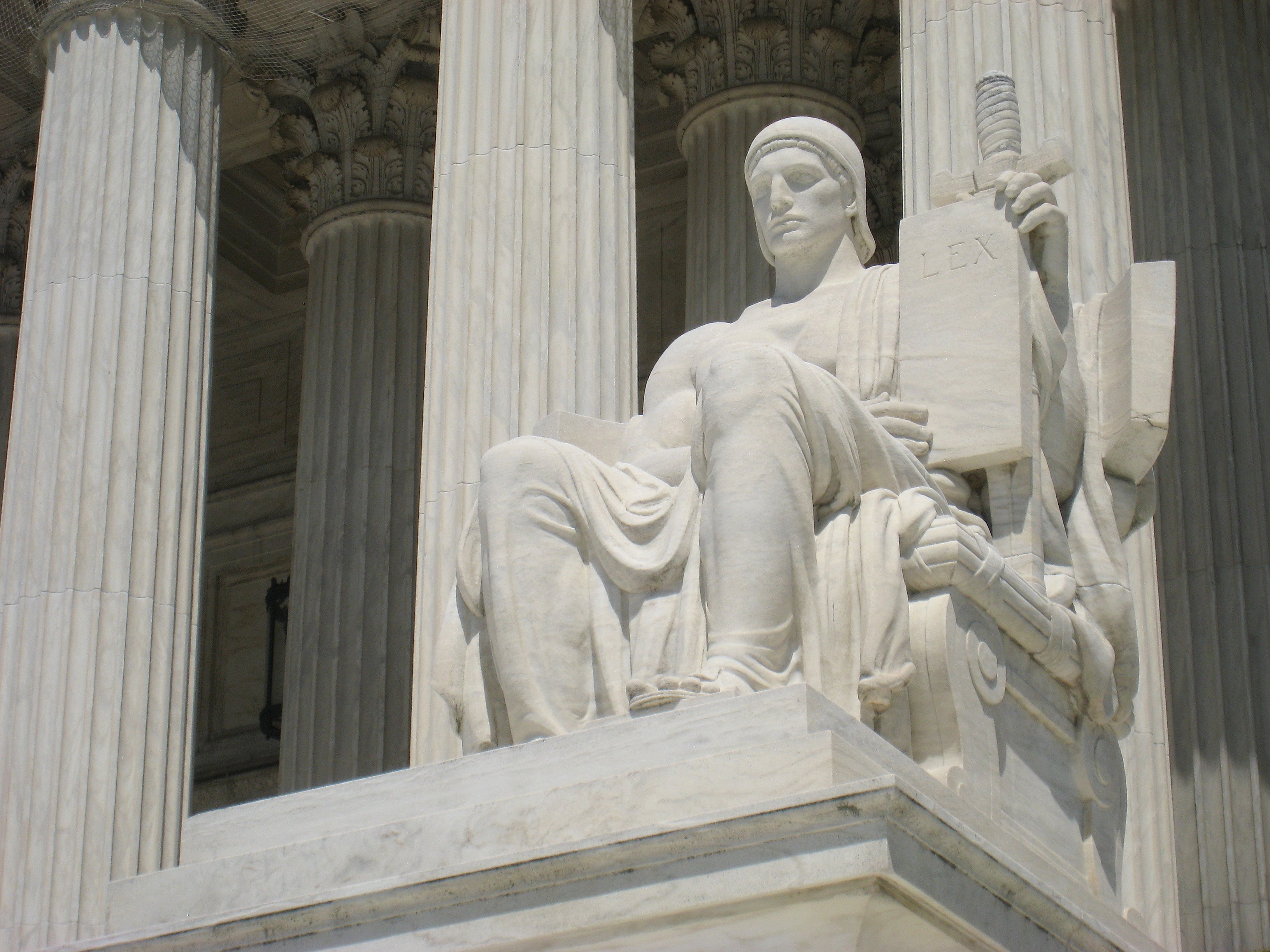 Photo of a white marble sculpture, sitting in front of the massive marble columns of the US Supreme Court building. The sculpture is of a seated man, wearing a draped robe and a head covering, and holding a sword in his left hand. His right right arm is across his lap, and in both hands he steadies a stone tablet that reads, "LEX."