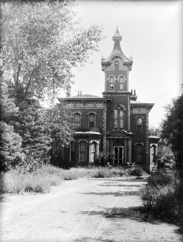 Photo of an old Victorian brick home, the Hindry house, which was at 5500 North Washington in Denver. The home has two floors and a tall tower providing a third floor and an elaborate scalloped roof. The windows are all tall with round arches at the top, and decorative brick frames. A dirt driveway leads up to the house, and narrow tire tracks are visible. Thick trees and shrubs line each side of the driveway and house. All of the curtains are closed and one window in the tower is broken.