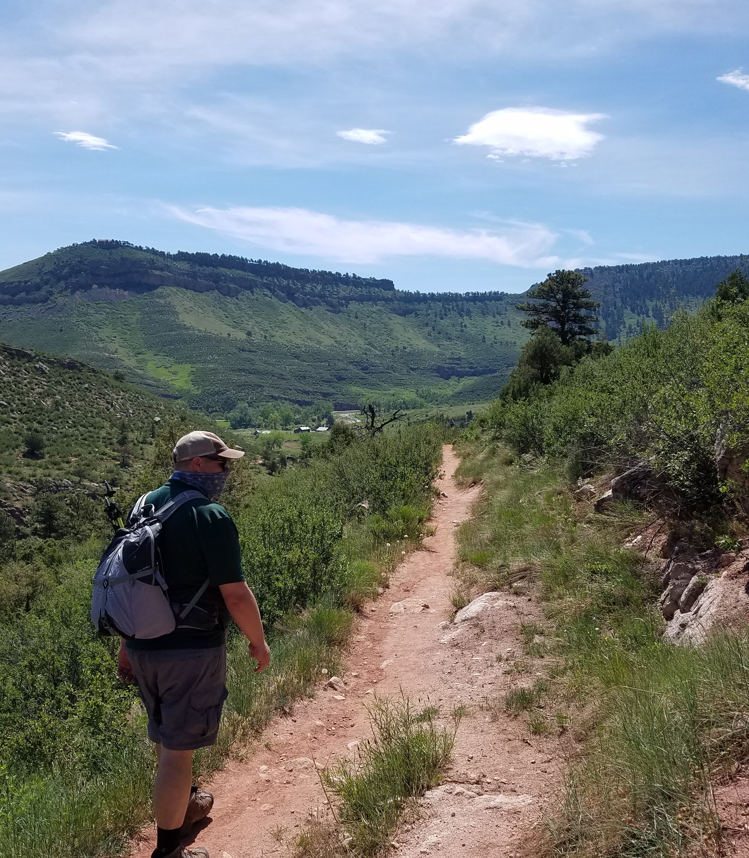 Photo of a lone hiker enjoying a sunny day on the trail. The green hillsides are visible in the background. In the foreground, a man wearing shorts, a t-shirt, a gray backpack, a baseball cap, and sun glasses is walking along the narrow dirt trail. He is also wearing a bandana over his mouth, in keeping with the Covid-19 protocols of the time.