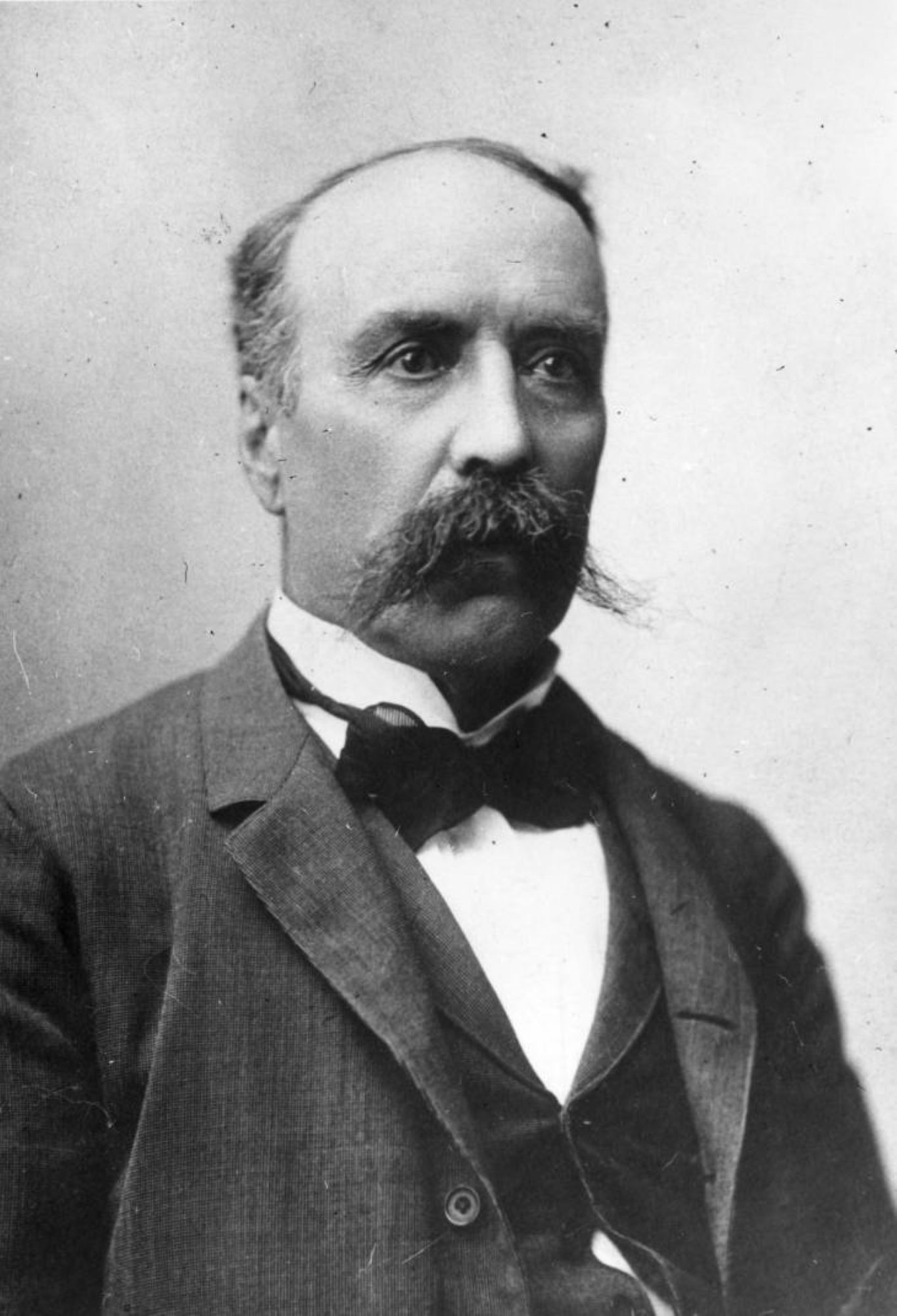 Photo of a middle-aged gentleman posing for a portrait. He is wearing a dark suit jacket and waistcoat over a white dress shirt with a black bowtie. He is balding, and his hair is short. He does have a very bushy handlebar-style moustache. He is not smiling, but rather looks straight ahead, off to the right of the image. The background of the photo is plain white.