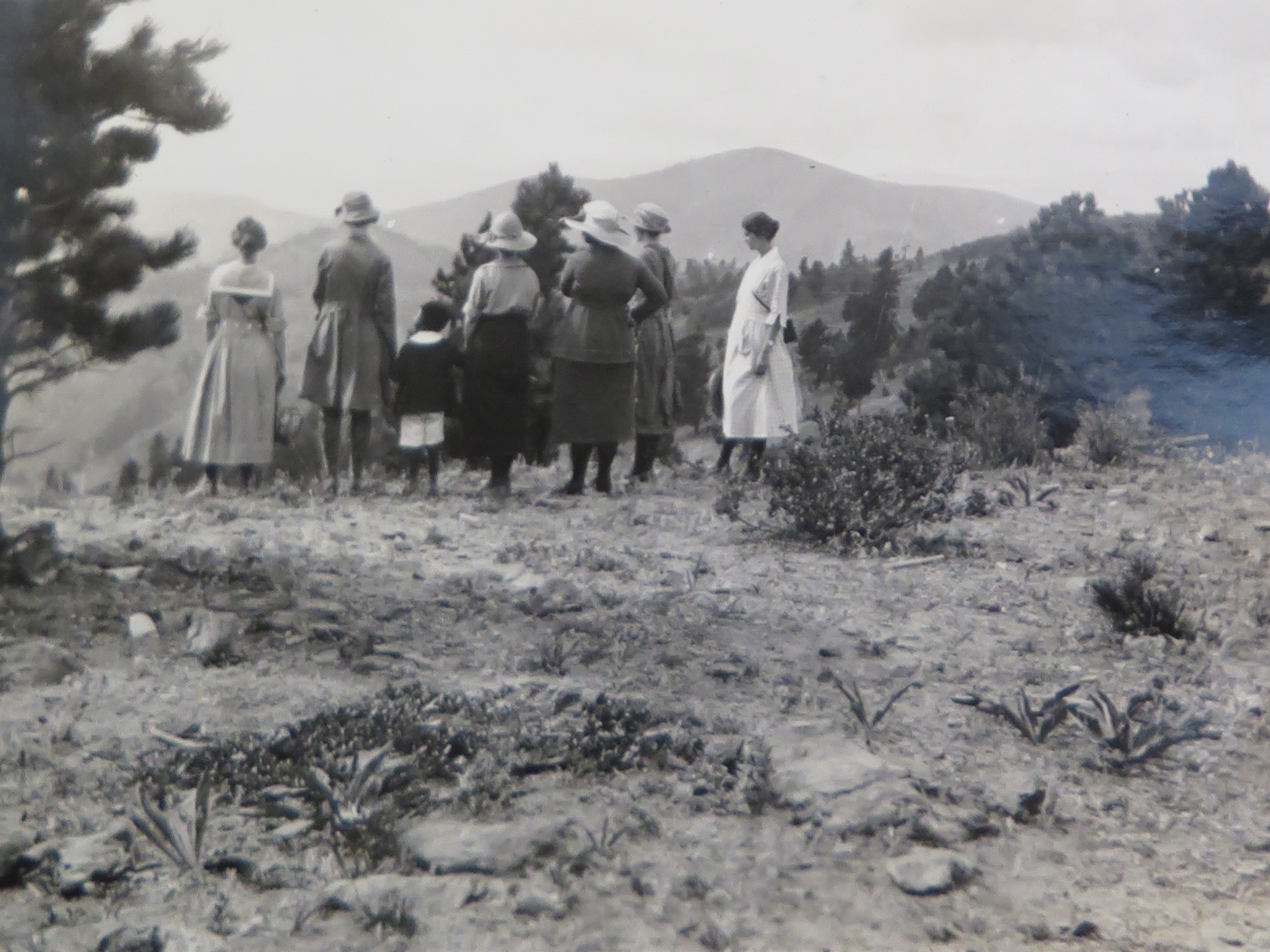 Photo of a group of women standing at the edge of a hill overlooking the mountains and valleys. Two of the women are holding the hand of a small Black boy, one on either side of him. He, too, is looking at the scenery. The women in this historic photo are all wearing dresses and most of them wear wide-brimmed hats. The young boy is wearing white shorts and a dark-colored shirt with a white collar.