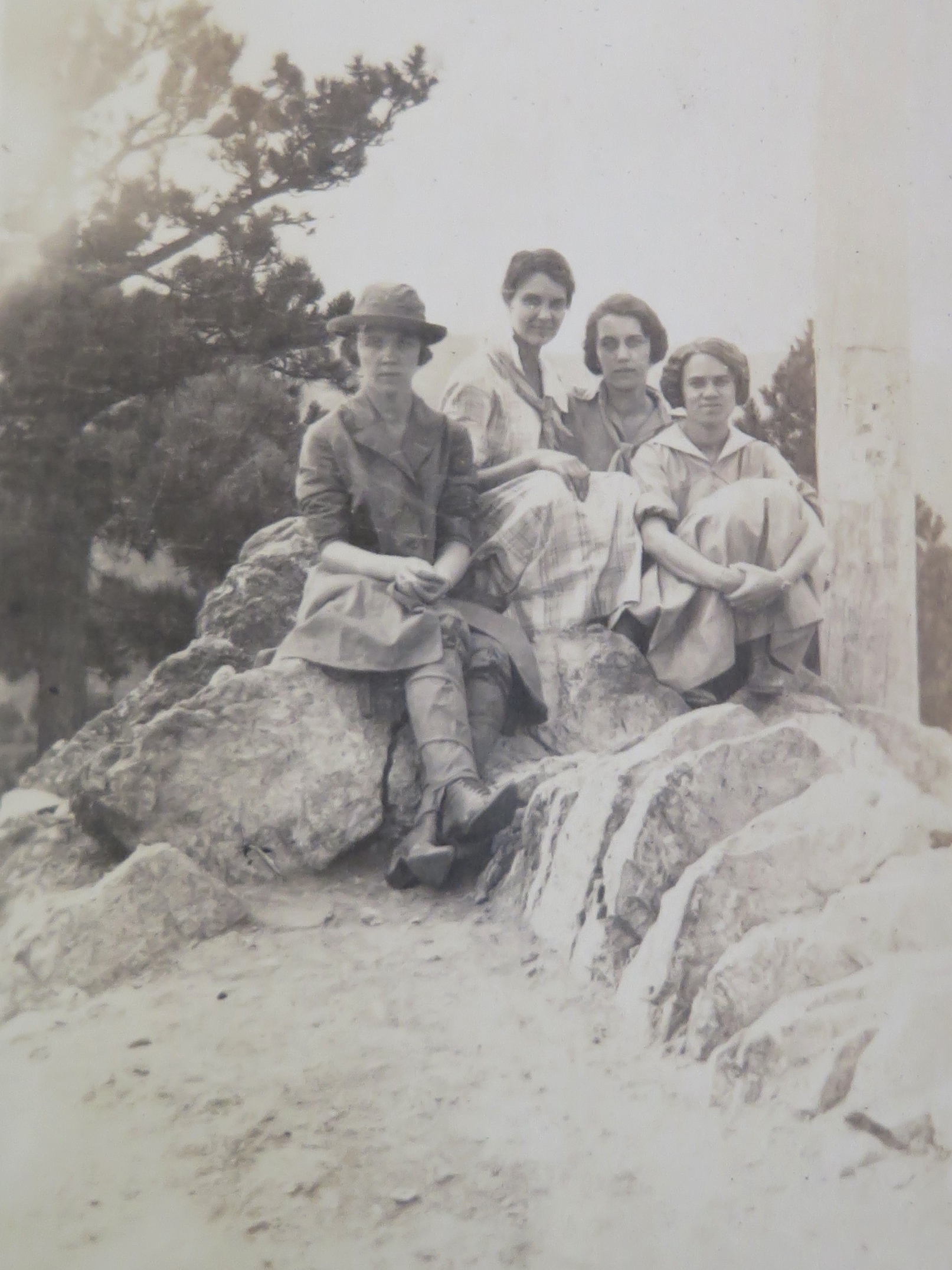 Photo of four women posing for a photo. The are sitting on some large boulders, and hold their skirts close to their knees as they sit. One woman, on the left, sits with her legs outstretched and her ankles crossed. She is wearing a skirt and tall boots, with a jacket and hat. The other 3 women don't have hats but wear skirts and have kerchiefs tied around their necks. All of the women either have short hair or have pulled their hair up on their heads.