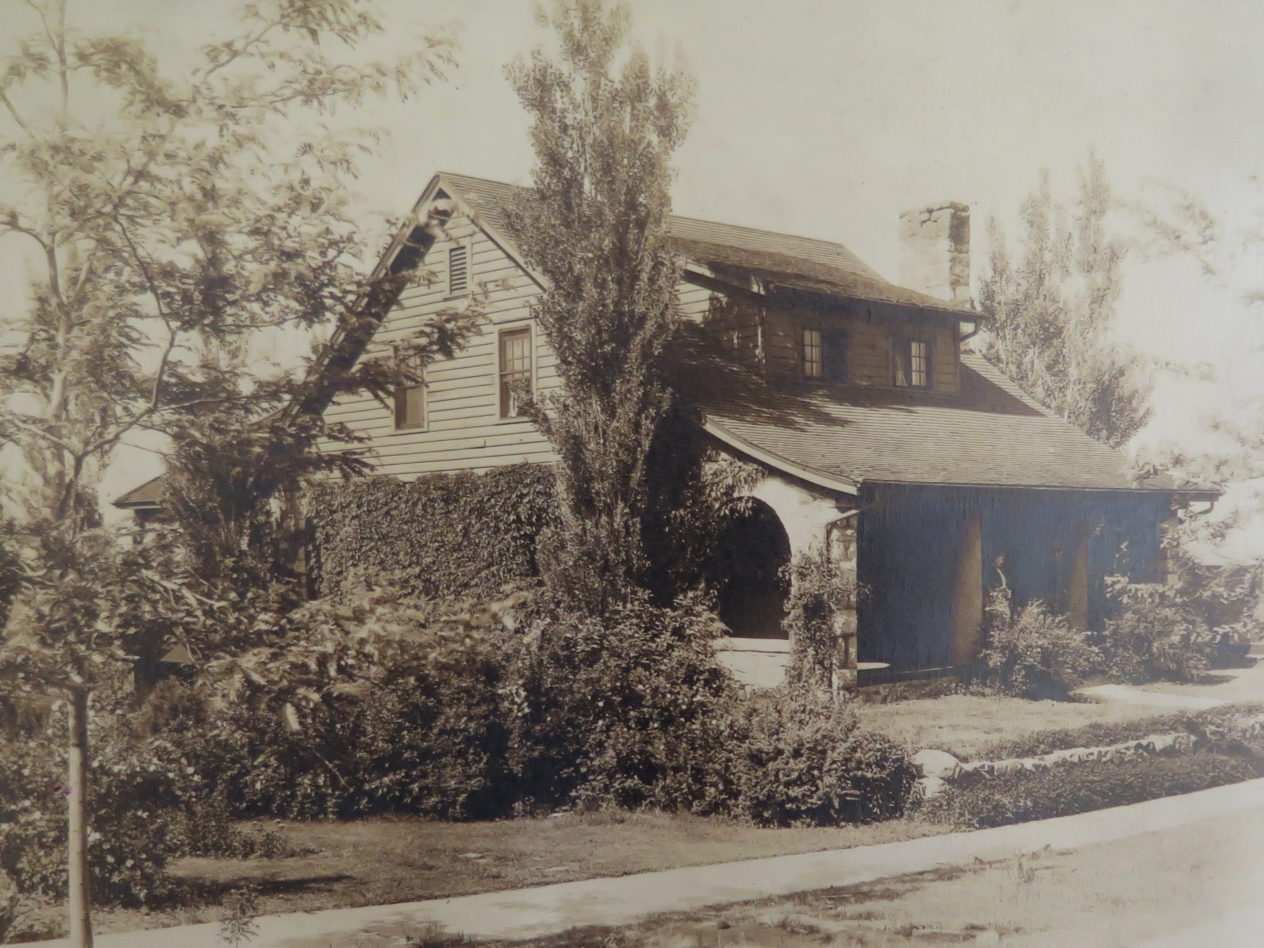 Photo of a vine-covered, two-story house with a shady front porch framed by arched openings. The wood-sided house is surrounded by shrubs, a few tall, thin trees, and a grassy lawn. A sidewalk stretches along the front of the property in this historic photo.
