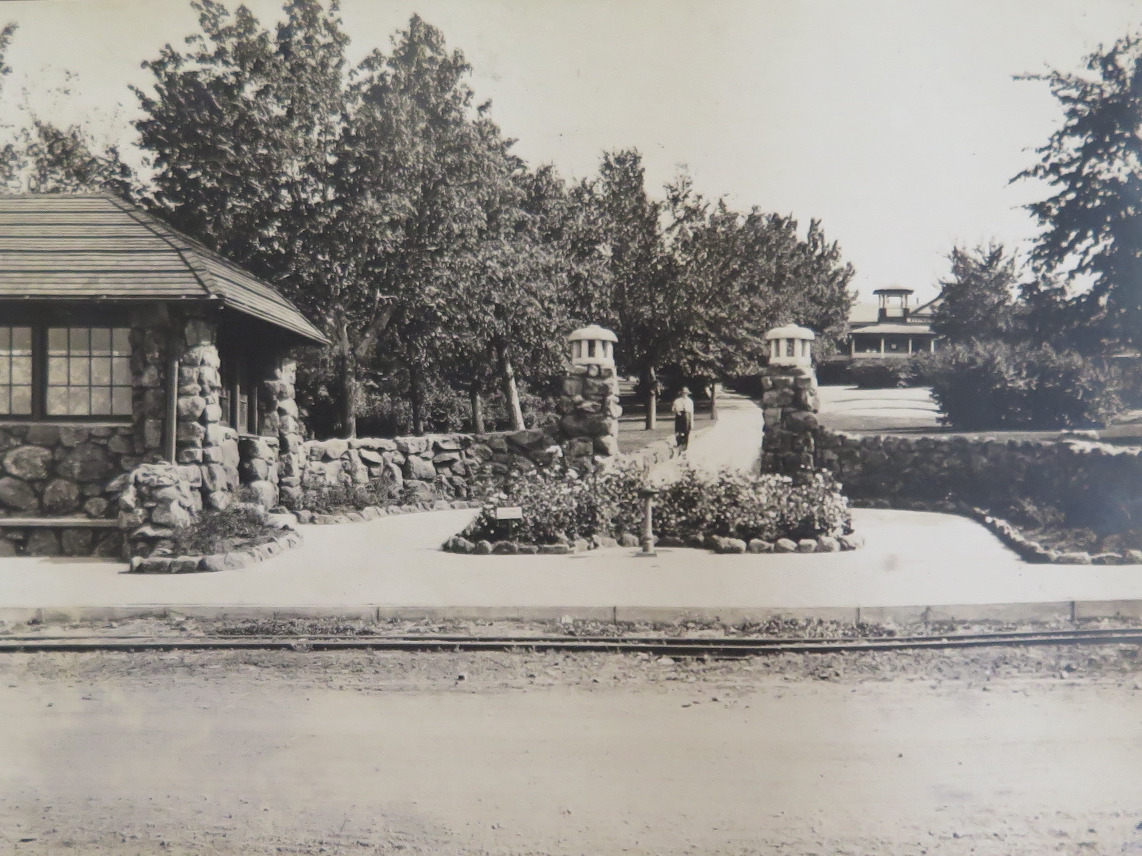 Photo of the entrance to a well-kept property. A small stone structure with large paned windows is on the left. From that building, a low stone wall emerges then curves outward, becoming an entrance marked by 2 pillars on each side. The wall continues off to the right side of the image. There is a small, low flower bed before the entrance, and someone can be seen along the walkway in the distance. A building with a tower and groomed lawns with tall trees lie beyond the walls.