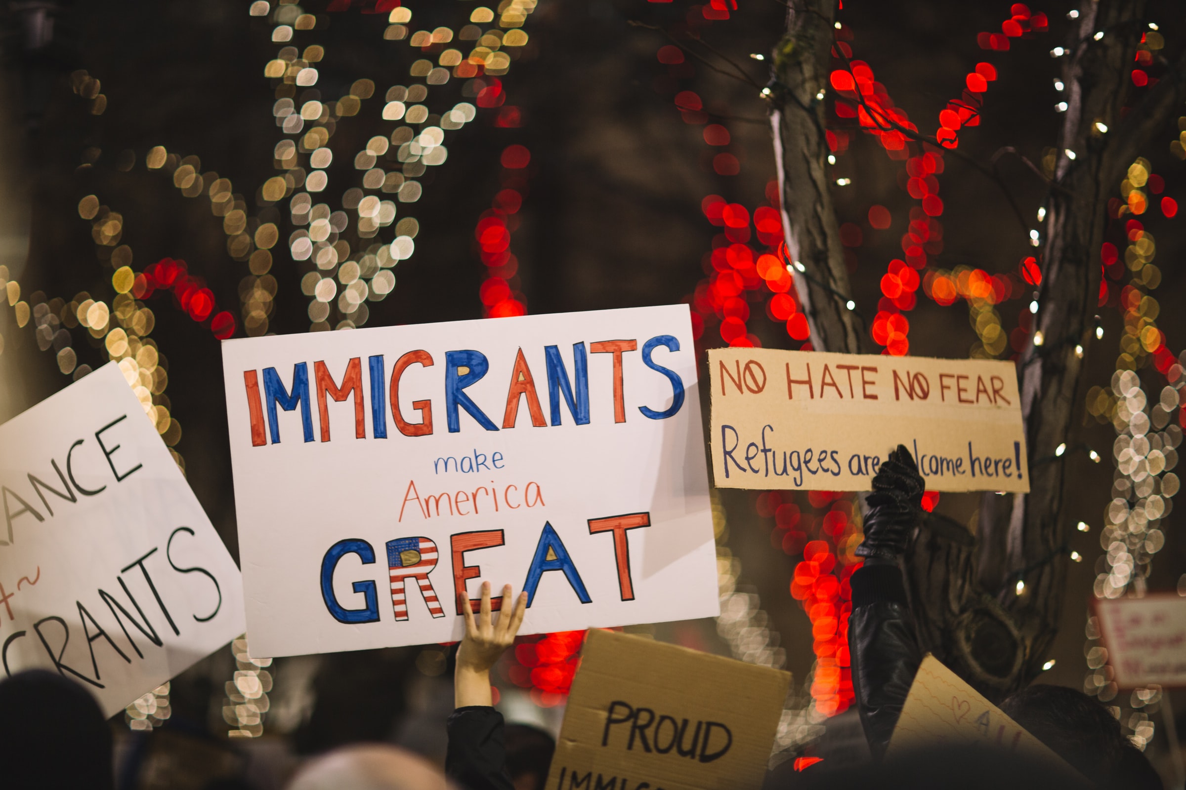 Photo of signs held up during a peaceful demonstration about Immigration in the US. While there are red and white lights in the trees in the background, in the foreground are signs that read, "Immigrants make America great" and "No hate, no fear, Refugees are welcome here!"