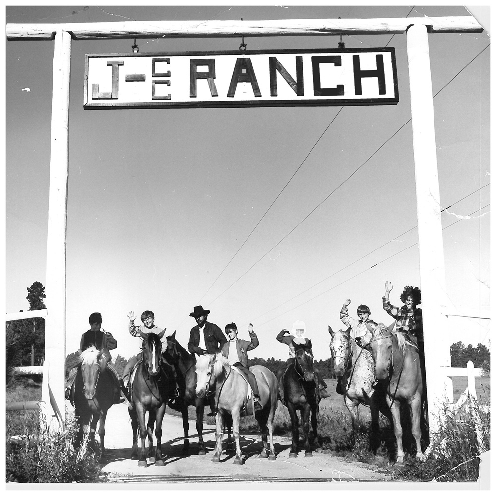 Photo of 6 young campers and a chaperone on horseback, standing below the front gates of the J Bar Double C Ranch. The campers are waving to the camera and smiling. All are wearing shirts with light jackets, and two wear hats. It appears to be a clear, warm day in this vintage photo.