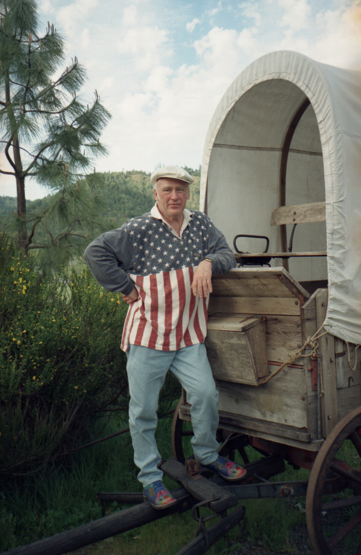 Photo of a man standing on the front hitch of a wooden covered wagon that is outdoors, surrounded by green shrubs and trees. He is wearing brightly colored shoes, blue jeans, and a white flat cap. His casual, rugby-style shirt represents the American flag: the top half of the shirt is blue with white stars, while the bottom half is red and white vertical stripes, and the collar is white. He is leaning his left elbow on the wagon and he is looking at the camera.