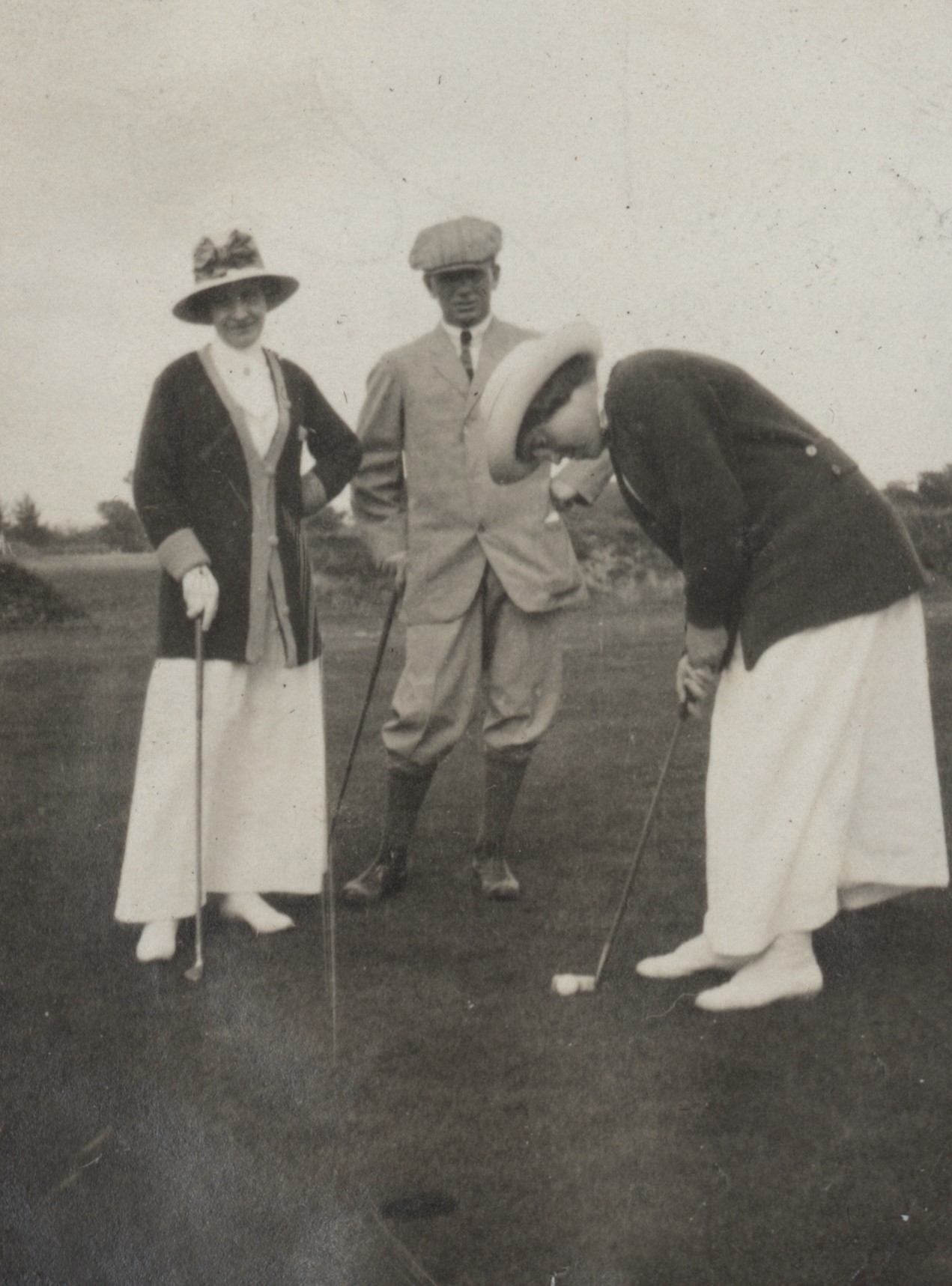 Photo of a man and two women playing golf. The woman on the right is getting ready to putt, and the hole is at the front of the photo. The other woman and the man are standing nearby, looking toward the camera. The man is wearing plus-fours golf attire with boots, a buttoned jacket, dress shirt and neck tie, and flat cap. The women wear long white skirts with long cardigans and wide-brimmed hats.